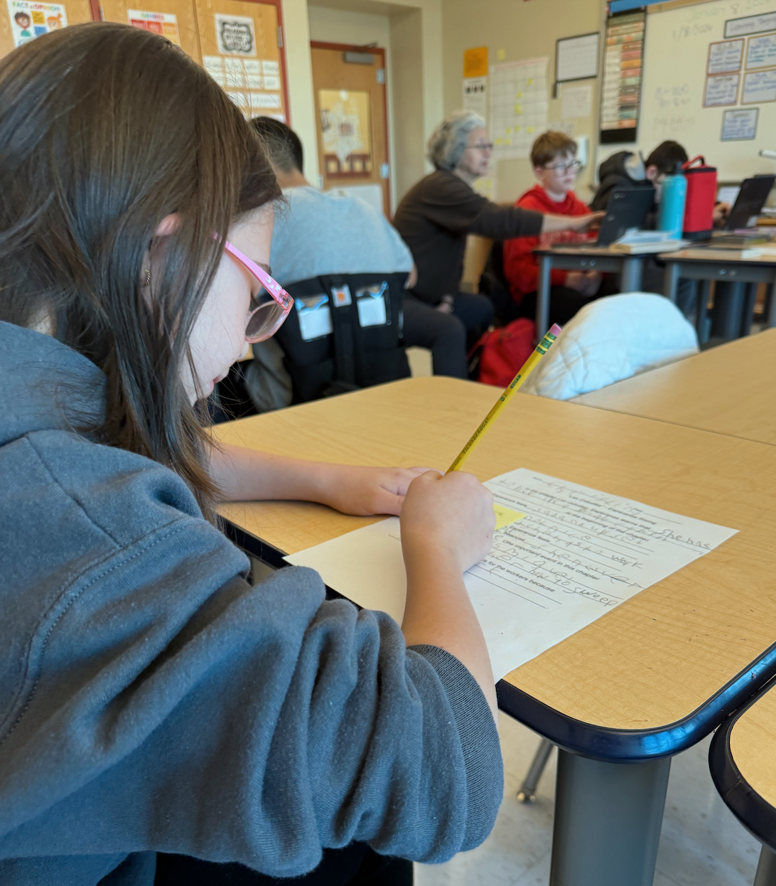 A young girl with glasses writing on a worksheet in a classroom, with other students and a teacher working on computers in the background.