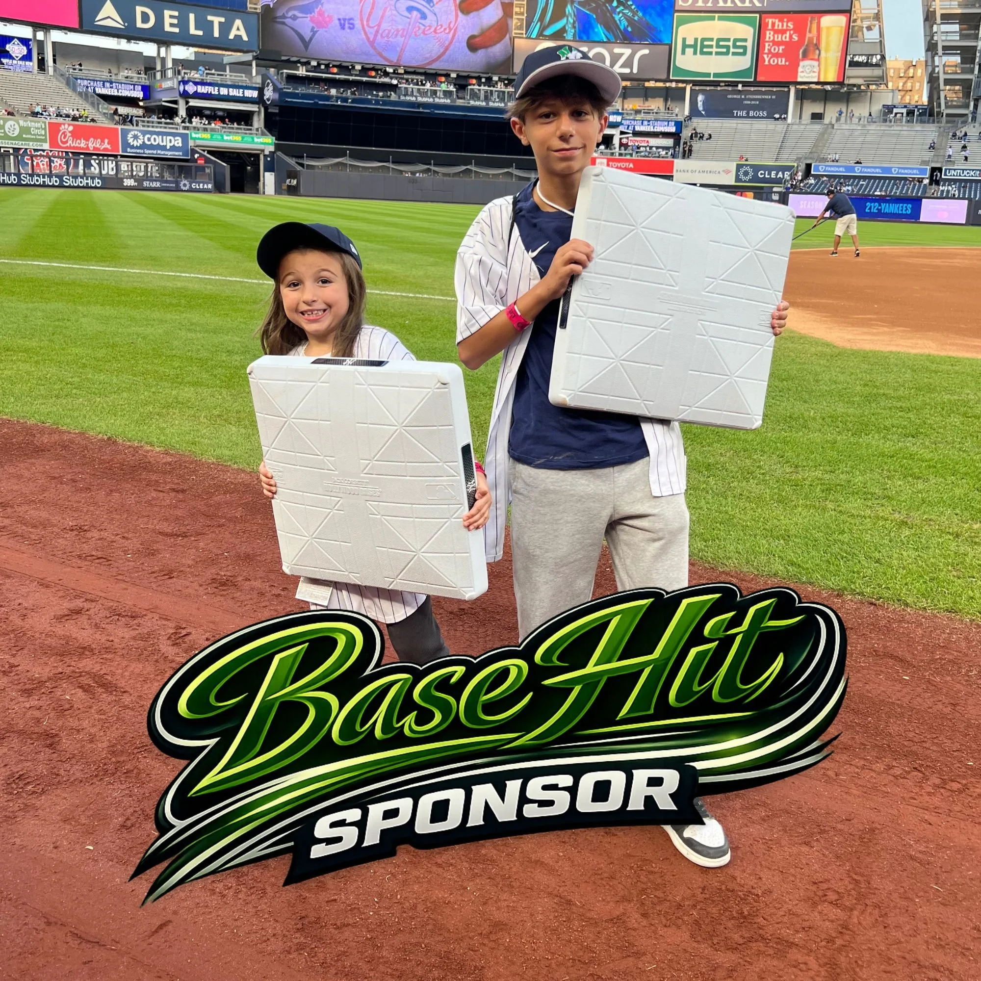 Two kids, a girl and a boy, standing on a baseball field holding white rectangular boxes, with the boy also holding a large sign that says 'Base Hit Sponsor'. The girl is smiling and wearing a baseball cap, the boy is wearing a gray cap, a baseball jersey, and gray shorts. The background shows the stadium with advertising banners and a player in the distance preparing to hit the baseball.