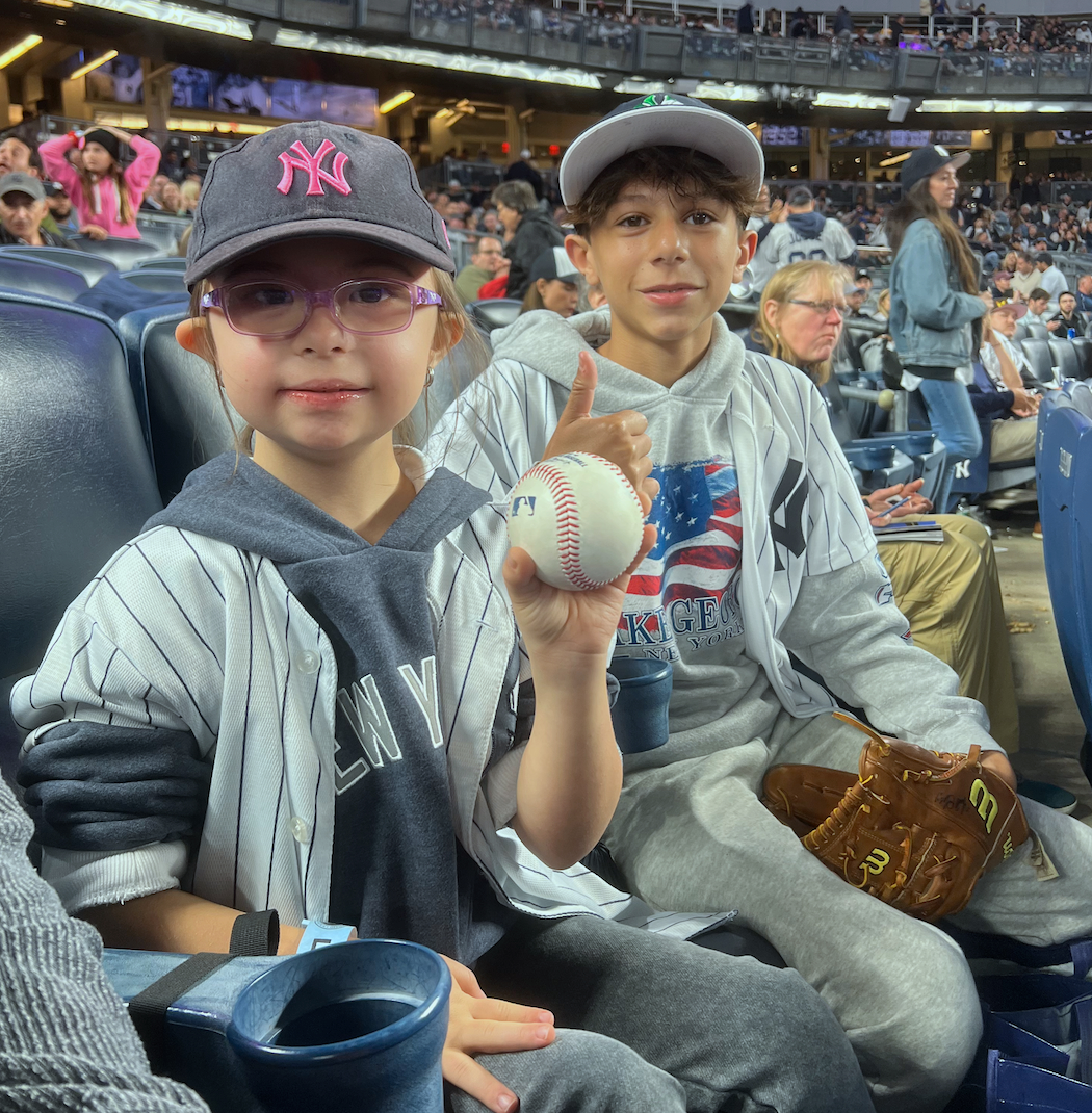 Two children sitting in a stadium, dressed in New York Yankees baseball jerseys and hats. The girl is holding a baseball and making a thumbs-up gesture. The boy is holding a baseball glove. The stadium is crowded with spectators.