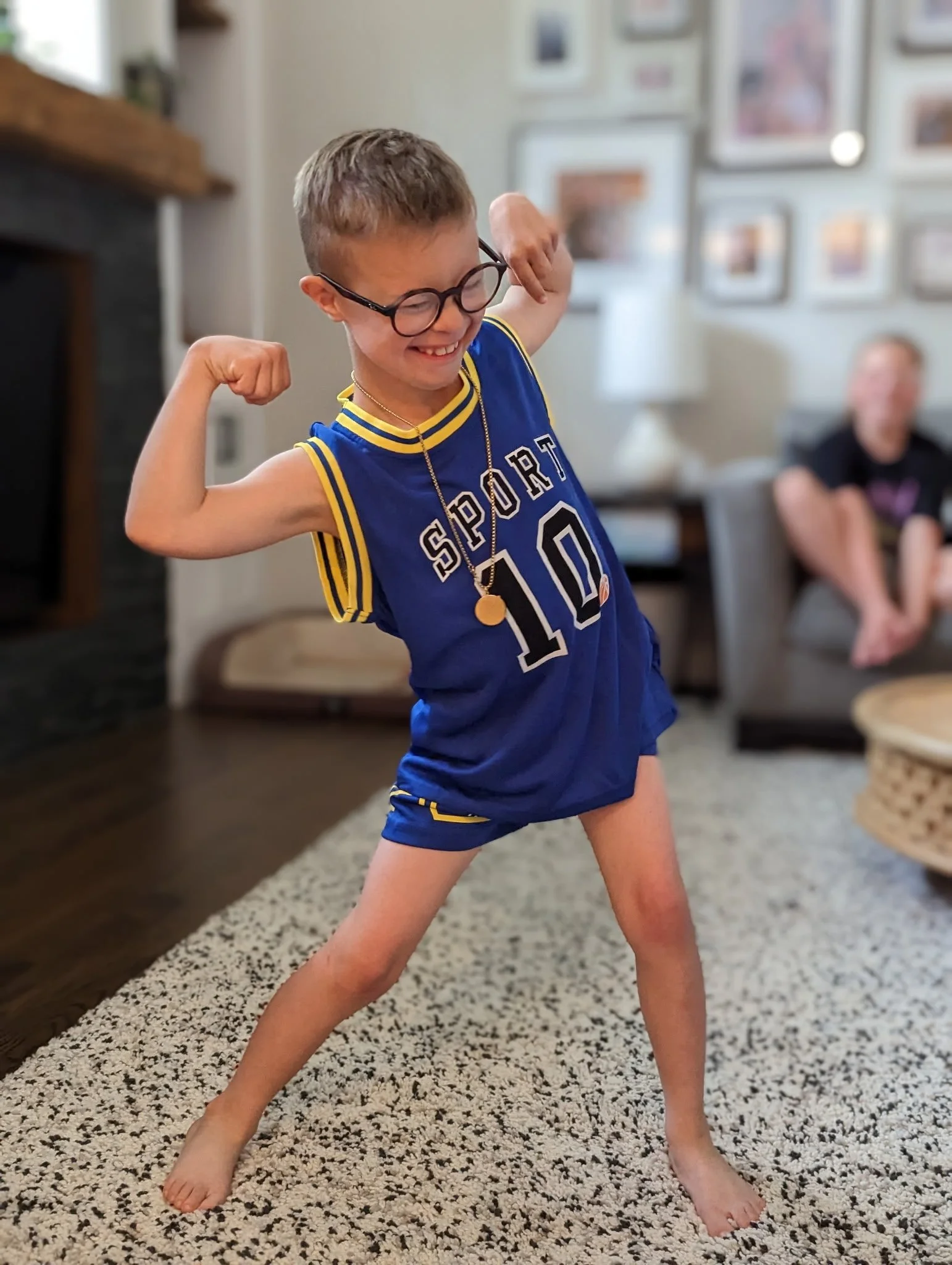 A young boy with glasses, wearing a blue sports jersey and shorts, posing with a flexed arm muscle and a big smile in a living room.