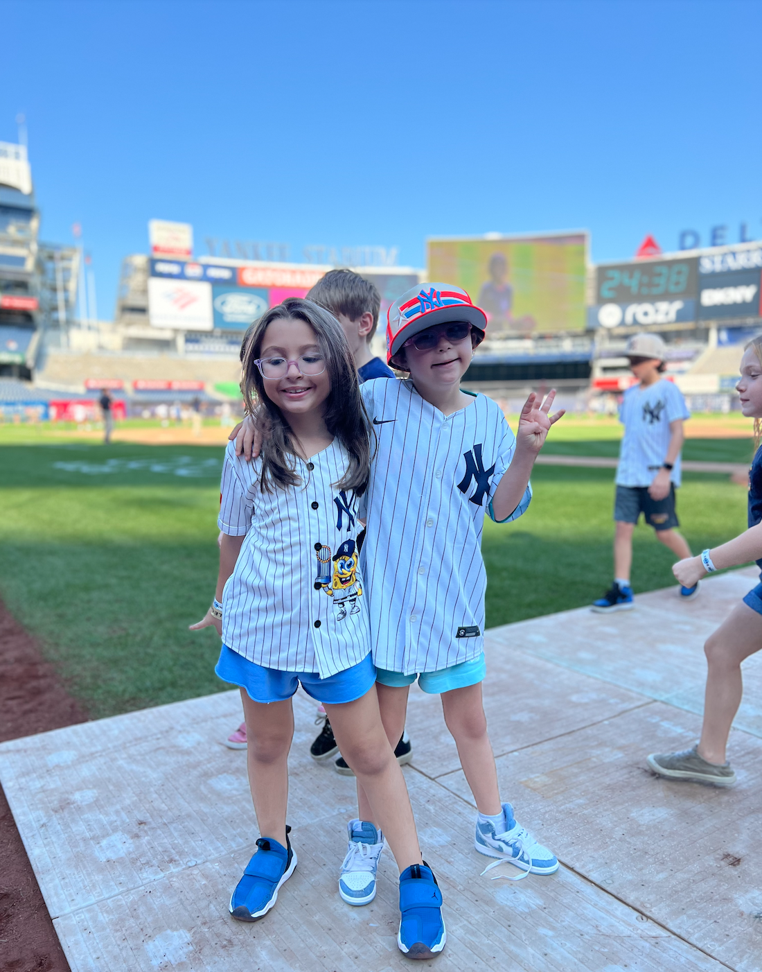 Two children wearing New York Yankees baseball jerseys, smiling and posing together on a baseball field.