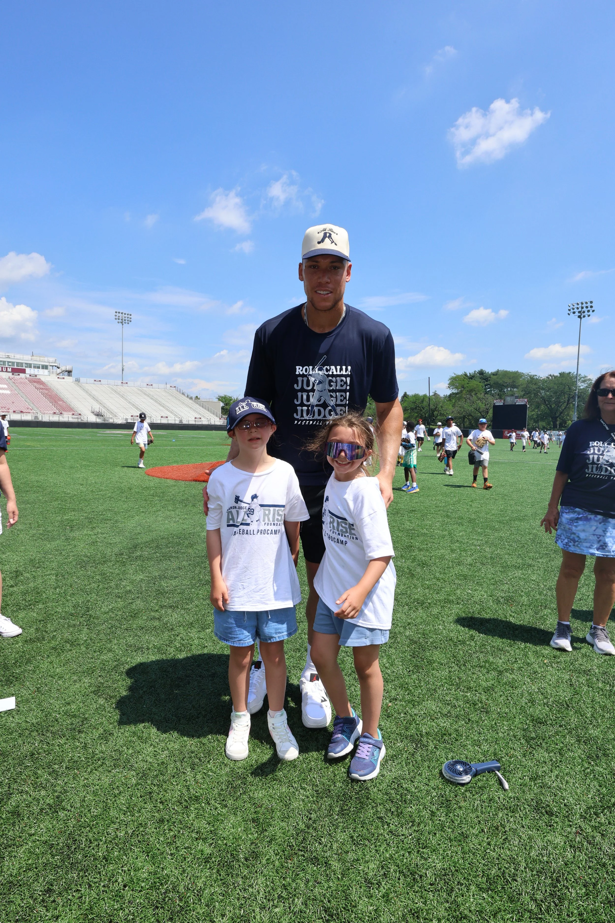 A man and two young girls standing on a grassy field at a baseball stadium during daytime. The man is wearing a navy blue T-shirt and a white cap, while the girls are in white T-shirts and sunglasses. Several people are seen in the background on the 
