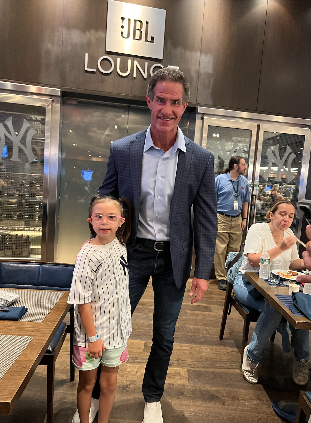 A man in a blue checked blazer and a young girl with glasses and a Yankees jersey standing together in a restaurant or lounge area, with a JBL Lounge sign in the background.