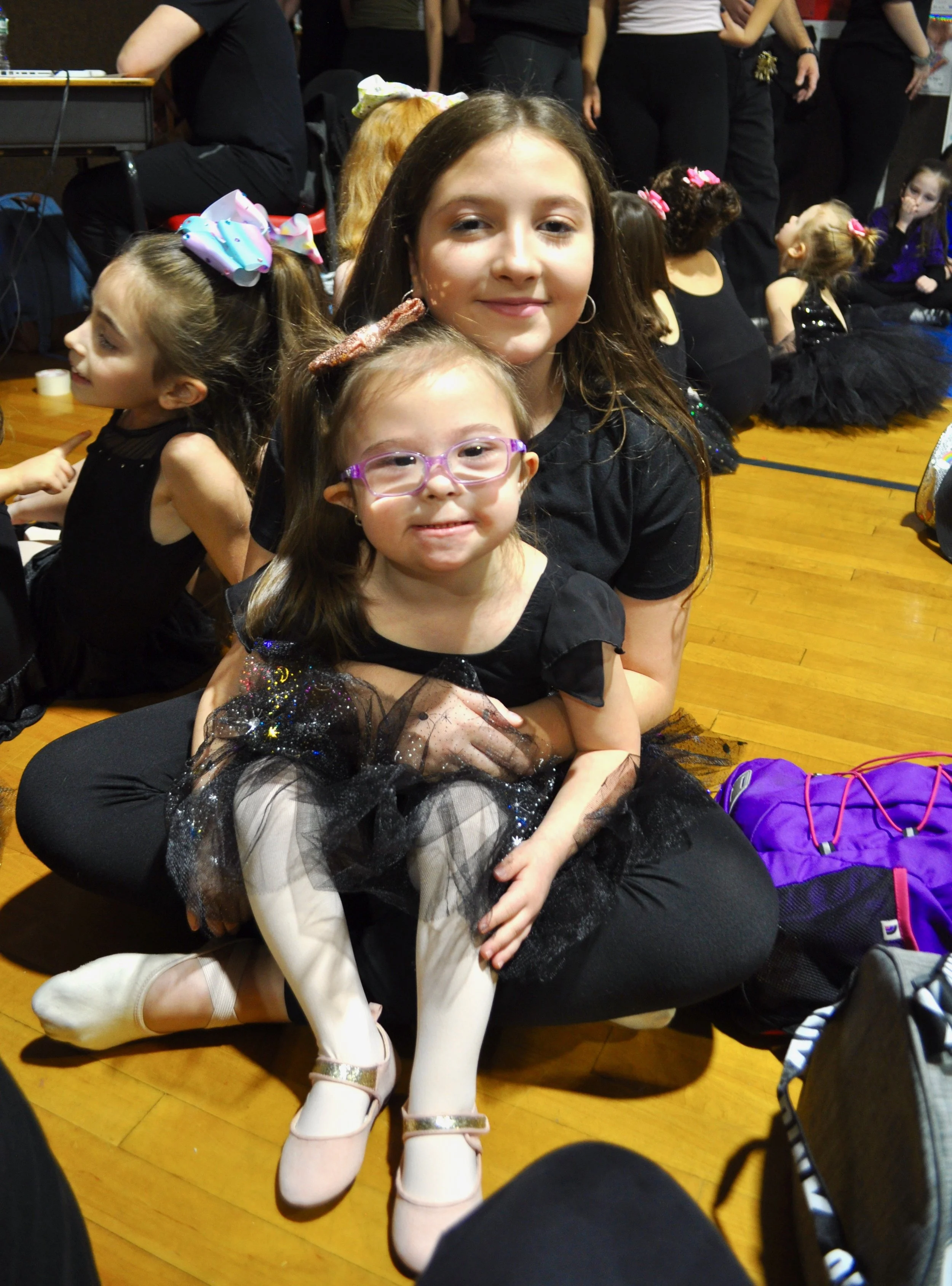 A young woman and a little girl sitting on the floor at a children's event or recital. They are dressed in black and have happy expressions. The background shows other children and adults, some dressed in costumes or dance attire.