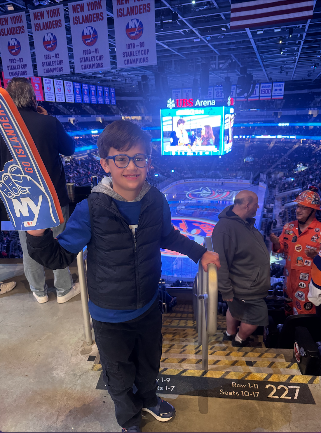 A young boy with glasses at an arena, holding a foam finger for the New York Islanders hockey team. The background shows the ice rink, banners hanging from the ceiling, and a large scoreboard. There are a few other spectators and a person dressed in 