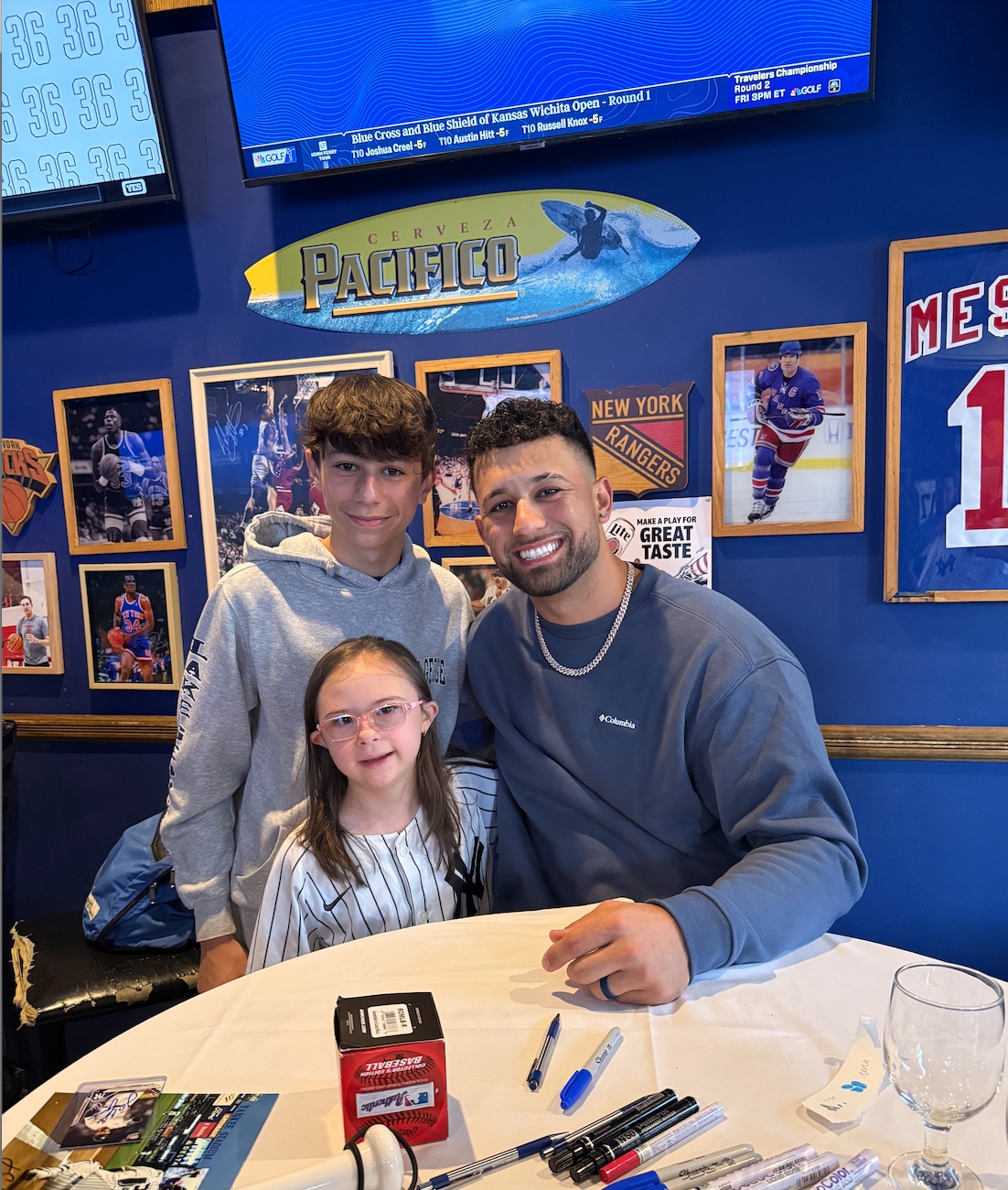 A man and two children sitting at a table in a sports-themed restaurant. The man and the boy are smiling, and the girl has glasses. The background shows sports photos and memorabilia, including hockey and basketball. There is a TV screen on the wall 
