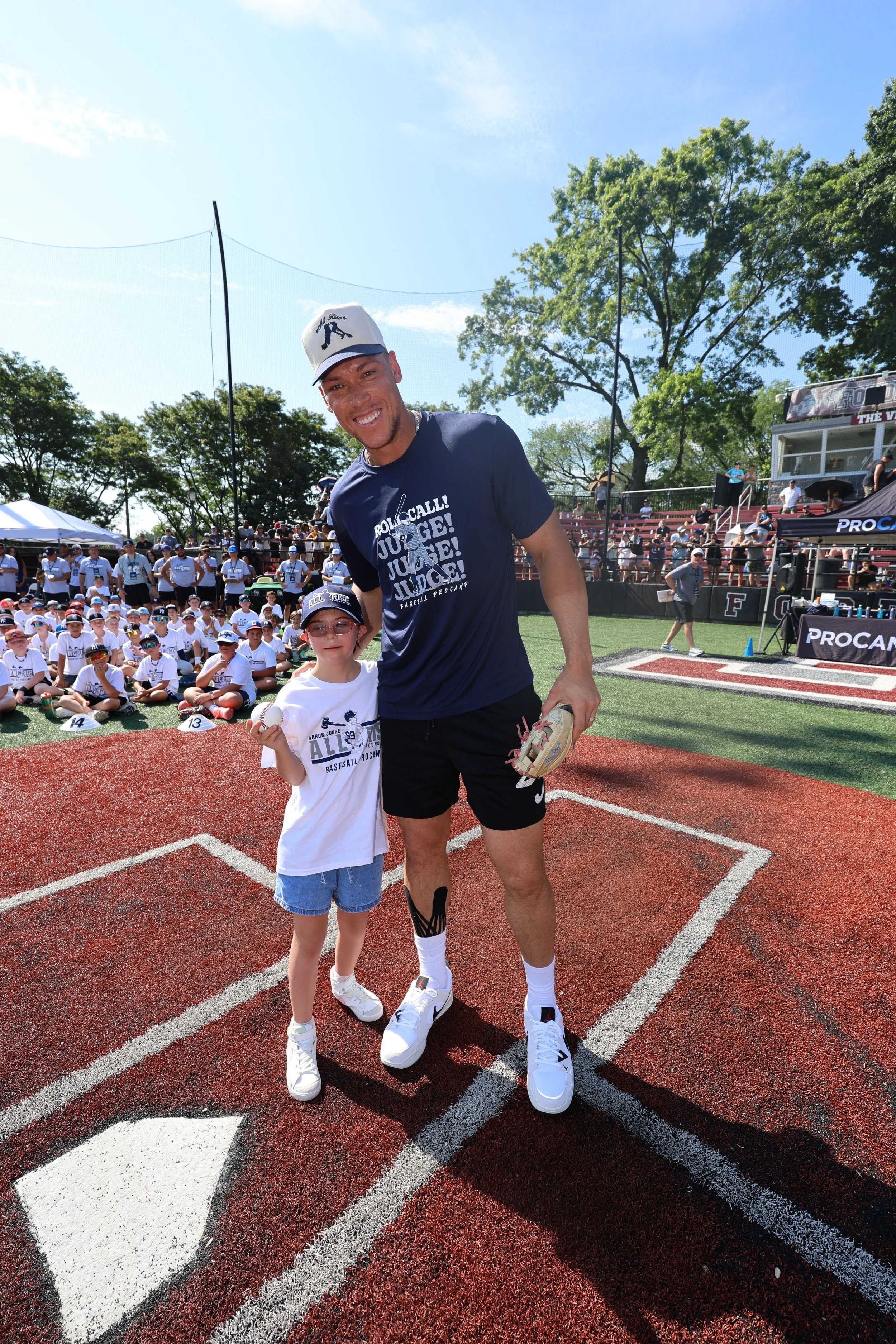 A young boy holding a baseball and a man with a baseball glove on a baseball field during a large event or game. The crowd is seated in the background, and the sky is clear and sunny.