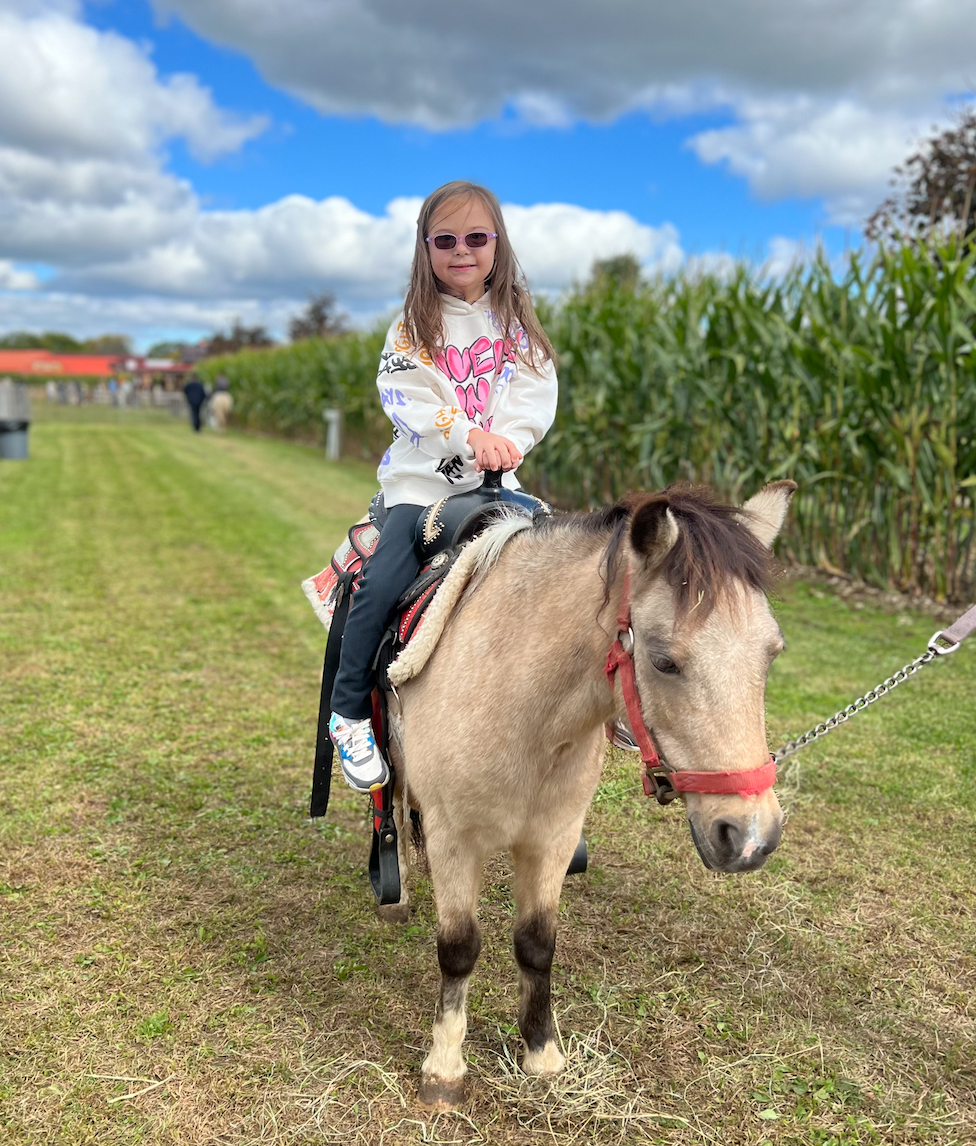 A young girl riding a small horse on a grassy field with a cornfield in the background. She is wearing sunglasses and a white hoodie with colorful writing, smiling at the camera.