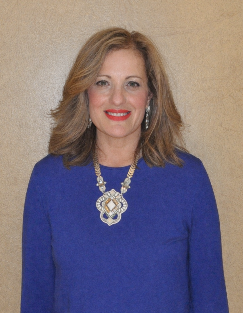 A woman with shoulder-length light brown hair, wearing a royal blue top, smiling, with a large statement necklace and earrings, standing in front of a plain beige wall.