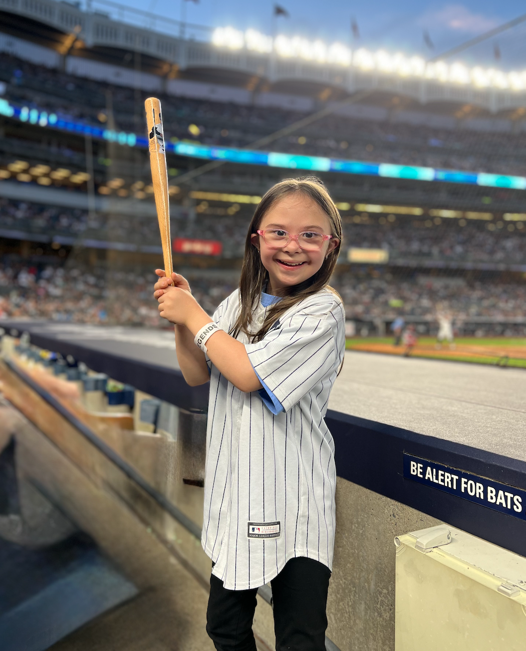 Young girl at a baseball stadium holding a bat, wearing a striped baseball jersey and glasses, smiling at the camera.