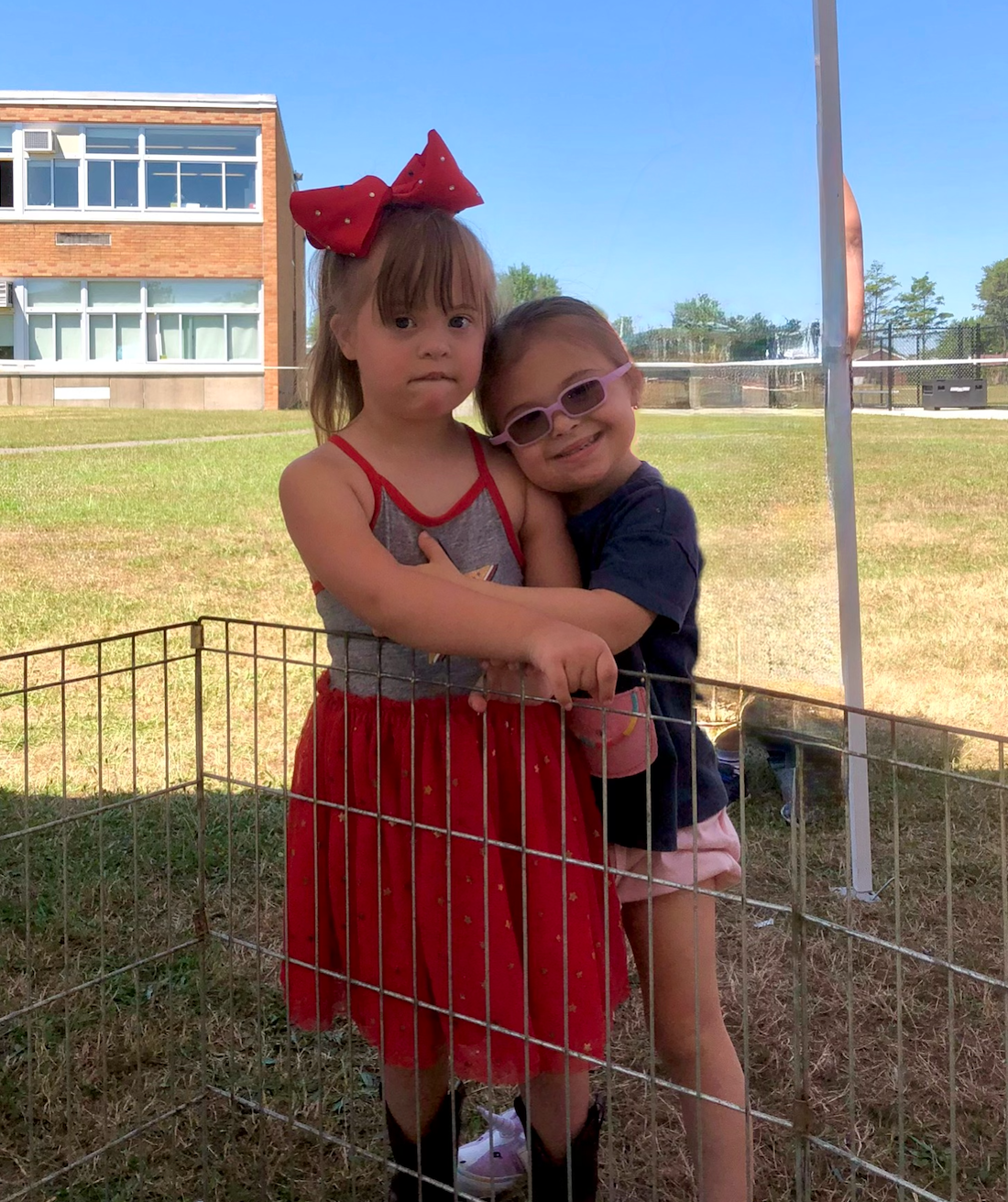 Two young girls hugging inside a metal playpen outside on a sunny day, with a school or campus building in the background.