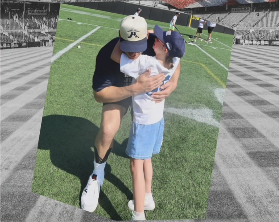 A man and a young girl hugging each other on a baseball field during a sunny day, with a few other people in the background practicing on the field.