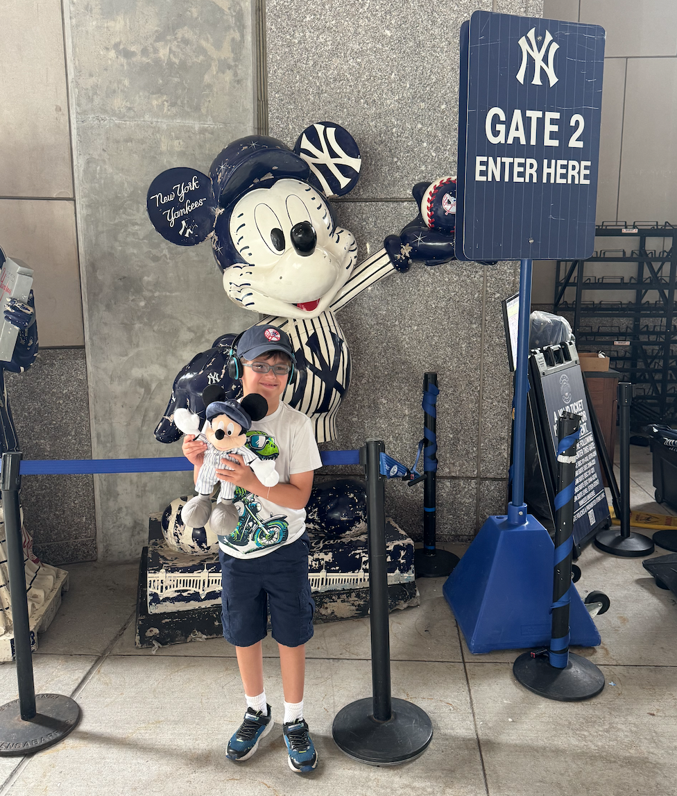 Child standing in front of a Mickey Mouse and Yankees-themed sculpture at an airport gate, holding a Mickey plush toy, with a sign indicating Gate 2 entrance.