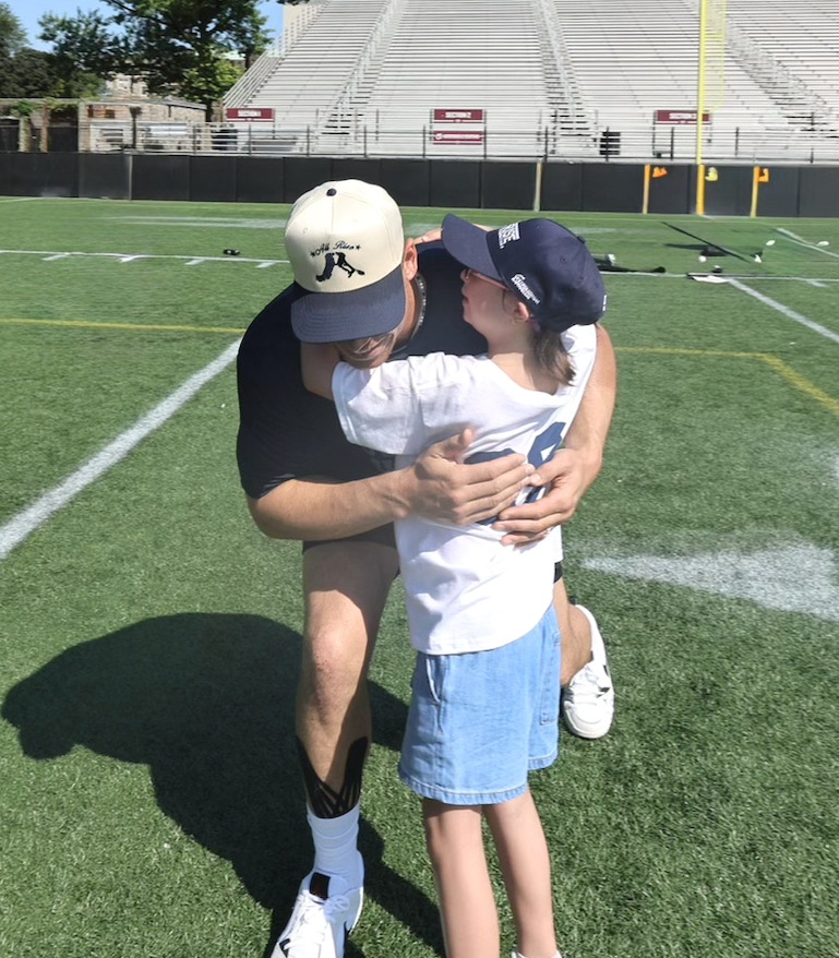 A man in athletic wear hugs a young boy on a football field, both wearing caps, with stadium seating and yard markers in the background.