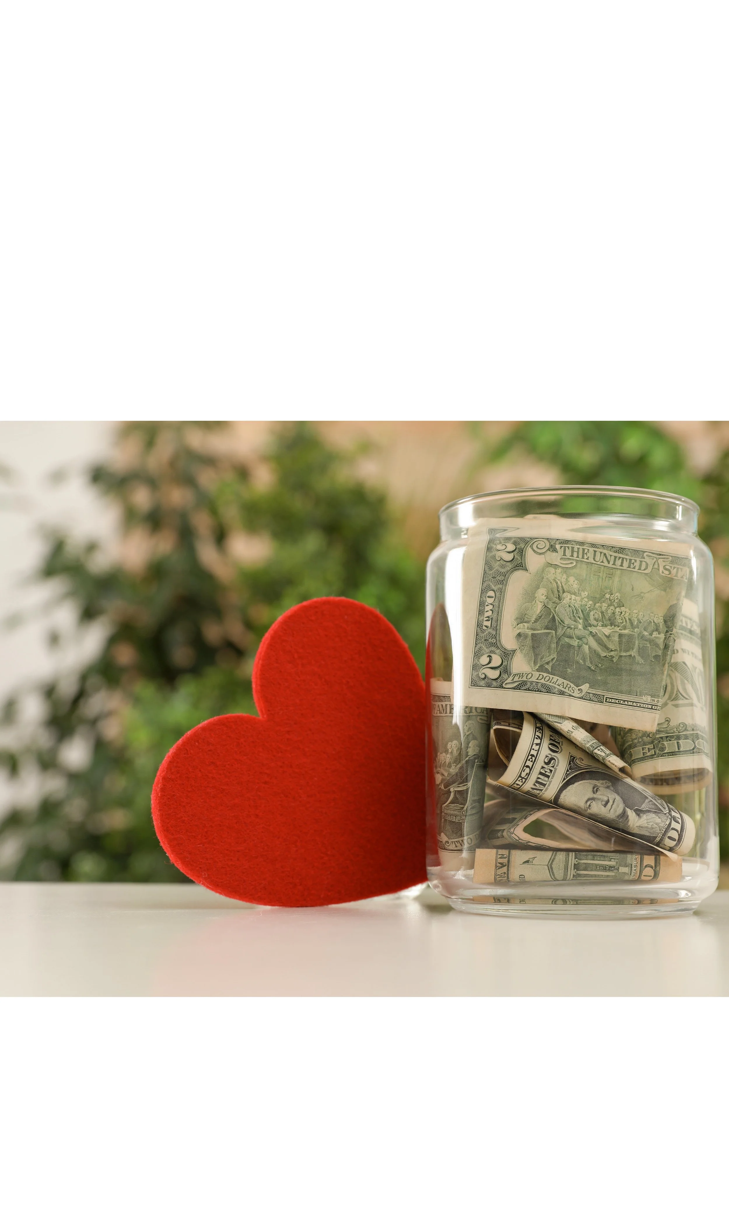 A glass jar filled with folded dollar bills next to a red felt heart on a white surface with a blurred green outdoor background.