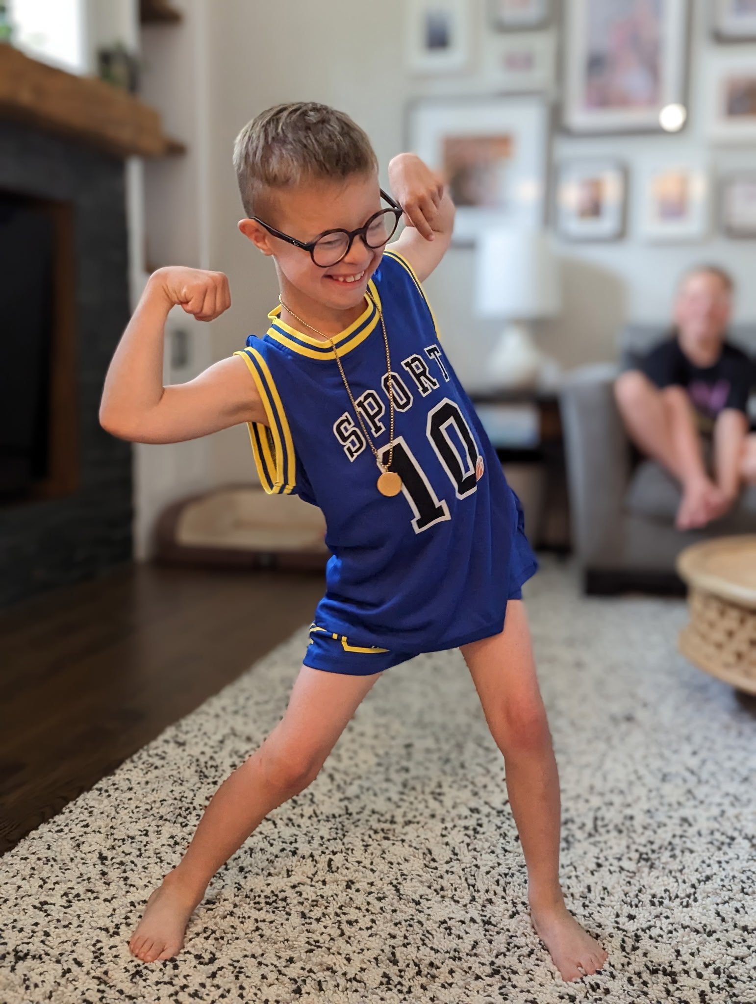 Child with glasses wearing a blue sports jersey and shorts, flexing muscles and smiling inside a living room.