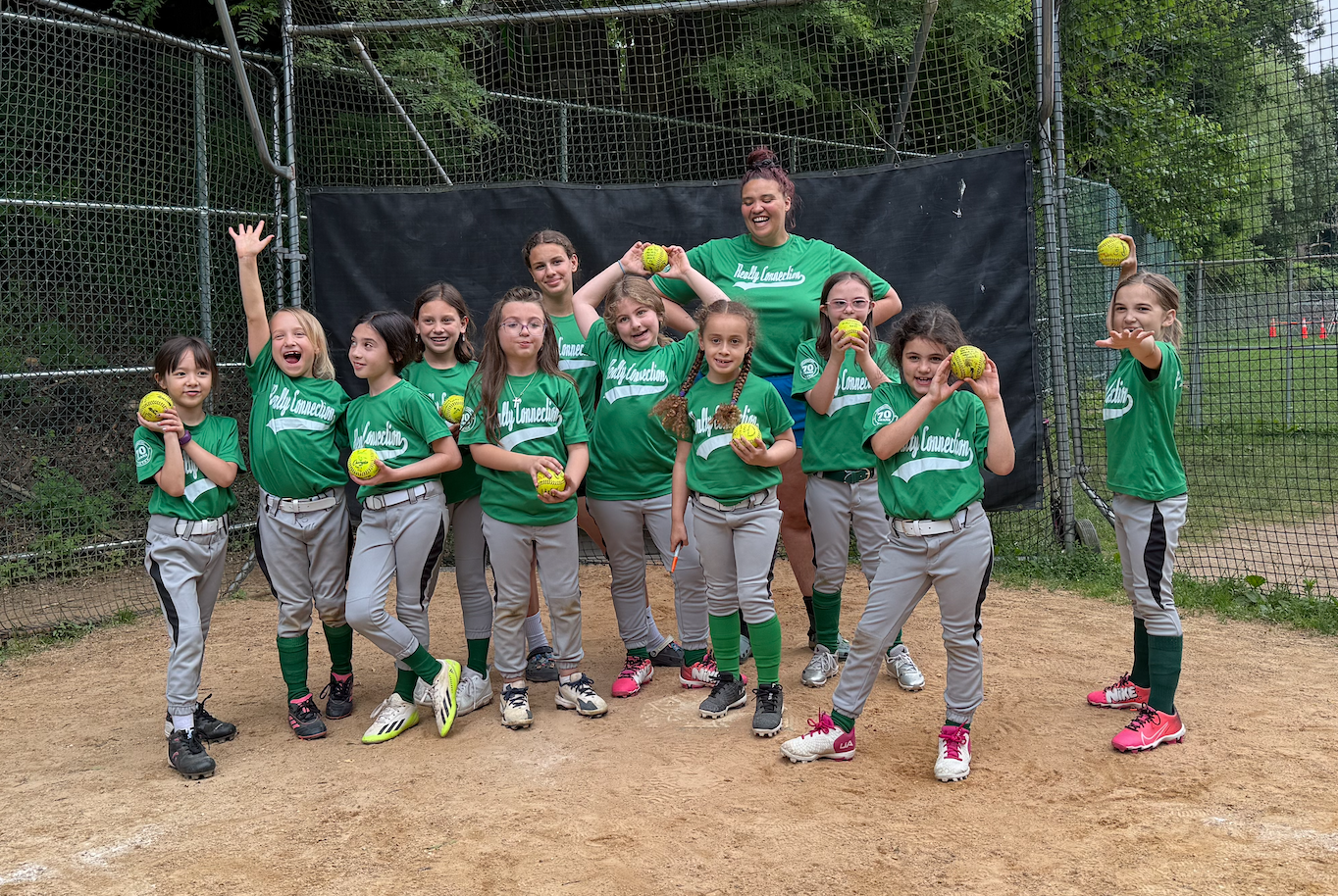 Young girls and a woman, presumably a coach or team member, pose for a team photo on a softball field. They are wearing matching green jerseys and gray pants, holding yellow softball gloves, with some holding softball mitts. The group appears happy and excited.