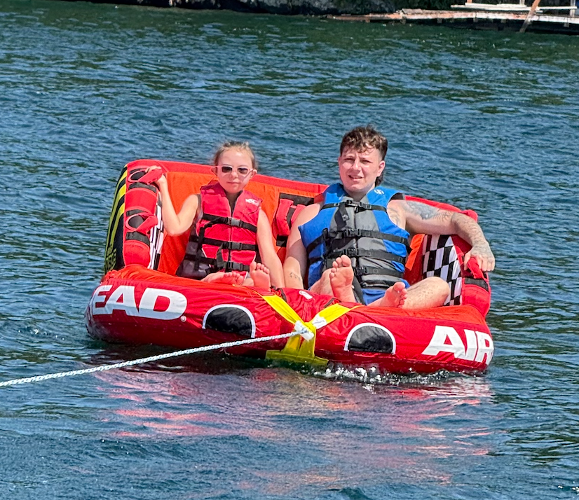 Two kids in life jackets sitting on a red inflatable towable inner tube on the water, holding onto the handle, being pulled by a boat.