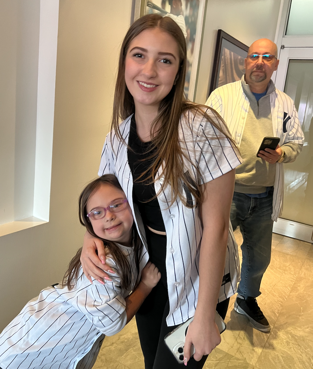 A young woman and a young girl, both wearing baseball jerseys, are posing together, with the girl hugging the woman. In the background, an older man is looking at his phone, and they are in an indoor setting with artwork on the walls and a glass door