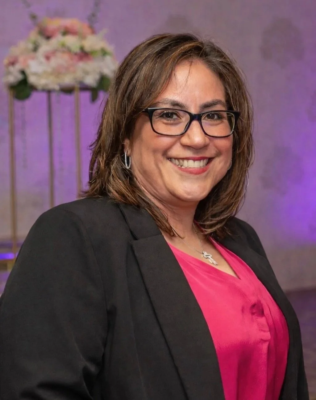 A woman with shoulder-length brown hair, wearing glasses, a black blazer, and a pink blouse, standing in front of a blurred background with purple lighting and a decorative flower arrangement.