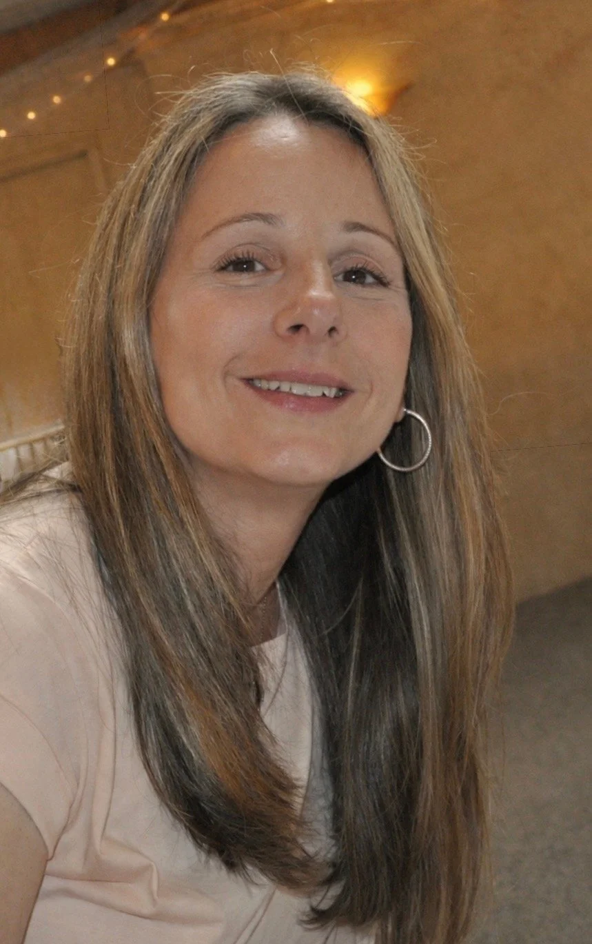 A woman with long brown hair smiling, wearing hoop earrings, in an indoor setting with warm lighting and a wooden background.