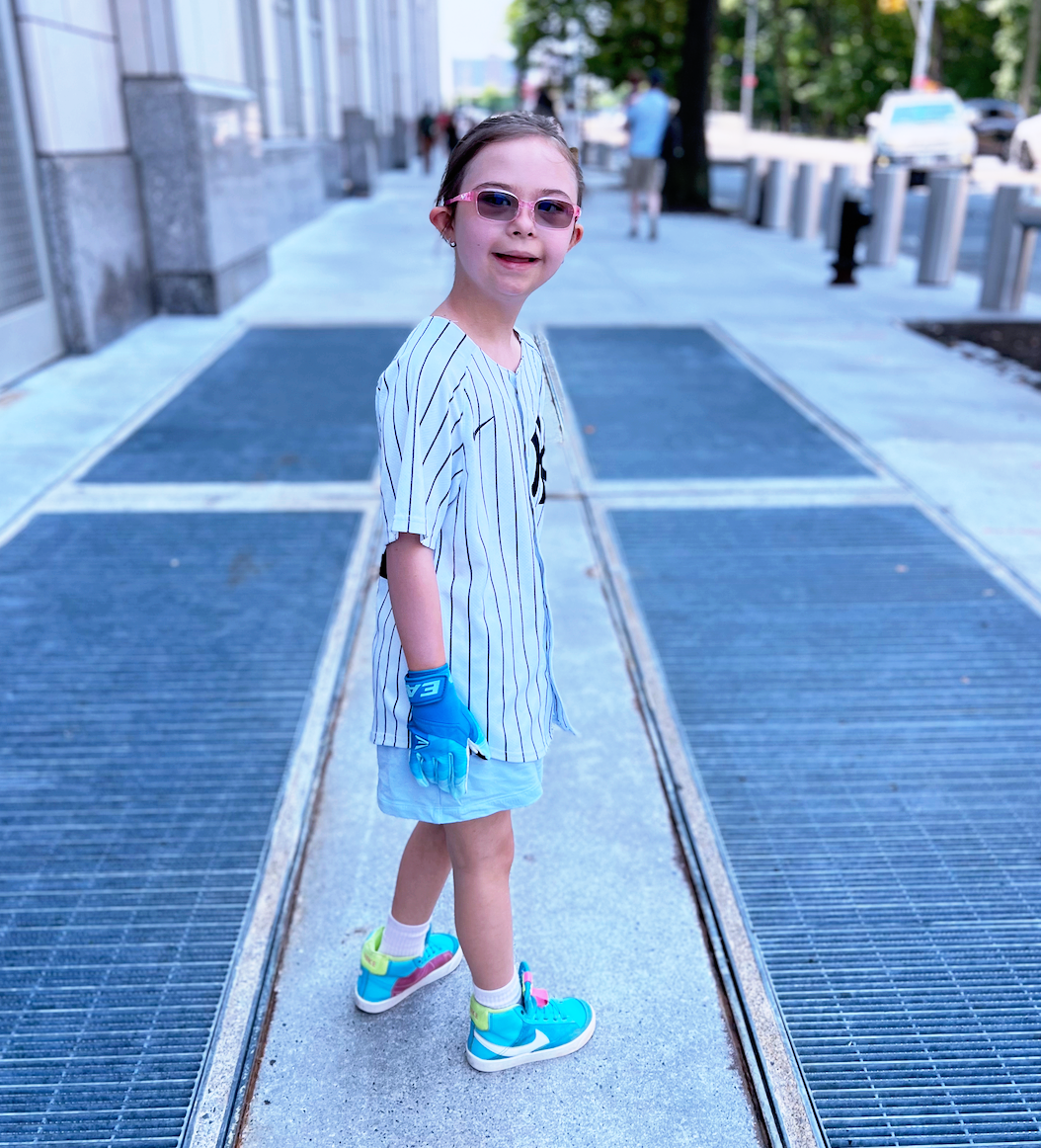 A young girl wearing sunglasses, a striped baseball jersey, shorts, and colorful sneakers standing on a sidewalk with metal grates, smiling at the camera.