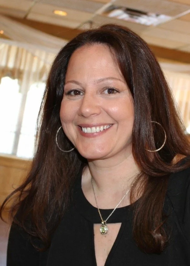 A woman with long brown hair, wearing hoop earrings and a necklace, smiling at the camera inside a room with large windows and curtains.