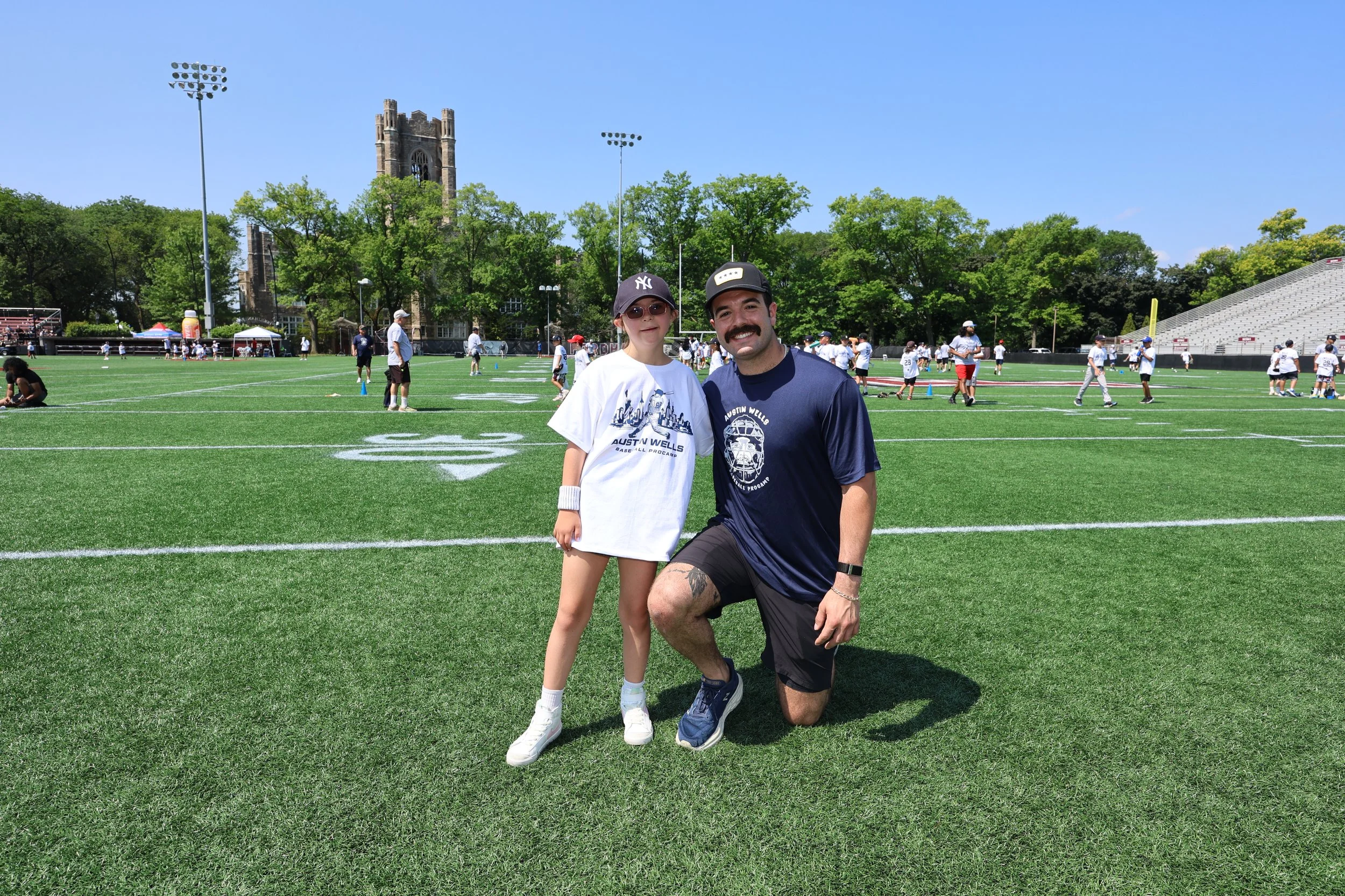 A person kneeling on one knee next to a girl on a sports field. The girl is wearing a white t-shirt, shorts, and a baseball cap. The person is wearing a navy blue t-shirt, black shorts, and a cap. In the background, children and adults are playing an