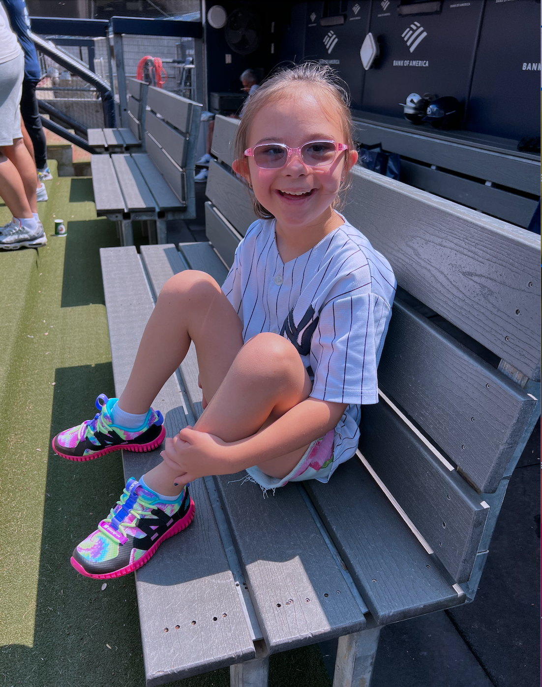 A young girl with blonde hair, pink glasses, and a big smile sitting on a wooden bench at a baseball stadium, wearing a baseball jersey, colorful sneakers, and shorts.