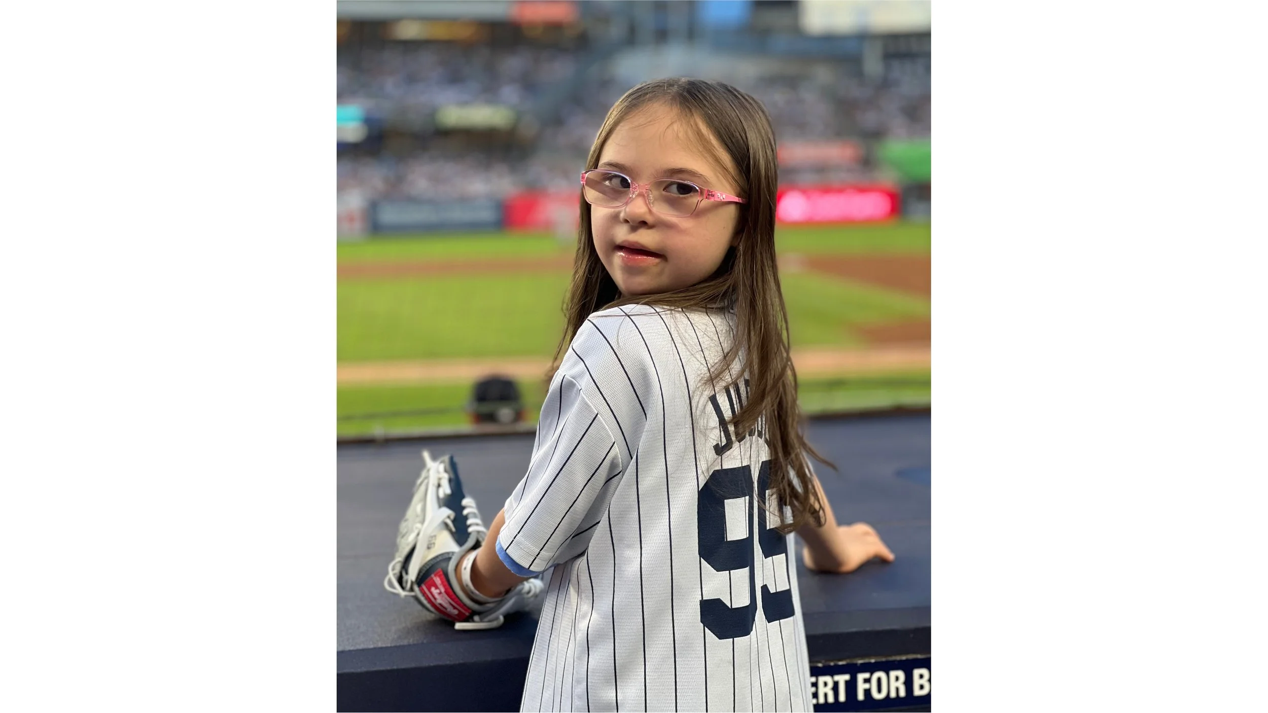 Young girl with long brown hair and pink glasses sitting on a stadium railing at a baseball game, wearing a white and navy pinstripe baseball jersey with the number 99 on the back, holding a baseball glove.