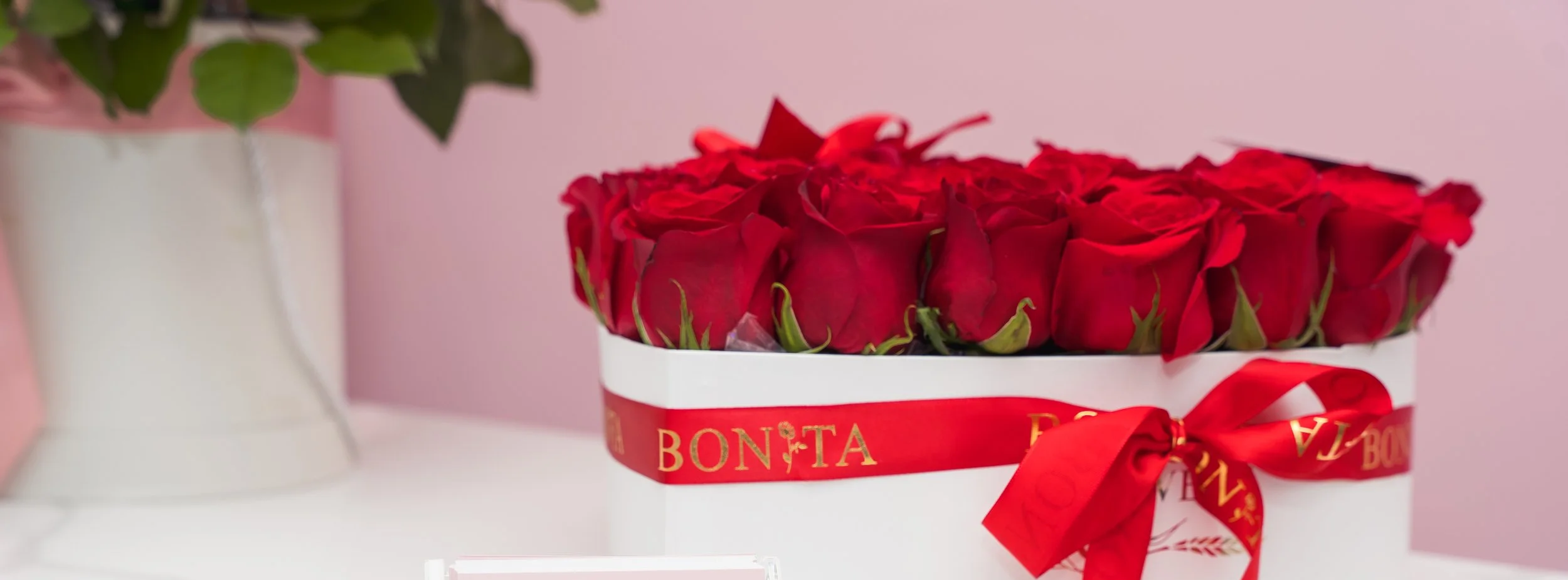 A white box filled with red roses wrapped with red ribbon that says 'BONITA' on it, placed on a white surface with a pink background.