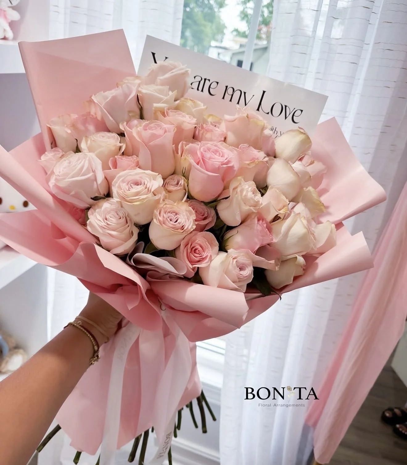 A person holding a bouquet of light pink and white roses wrapped in pink paper with a pink ribbon, with a card that says "You are my Love." The background shows a window with sheer white curtains.