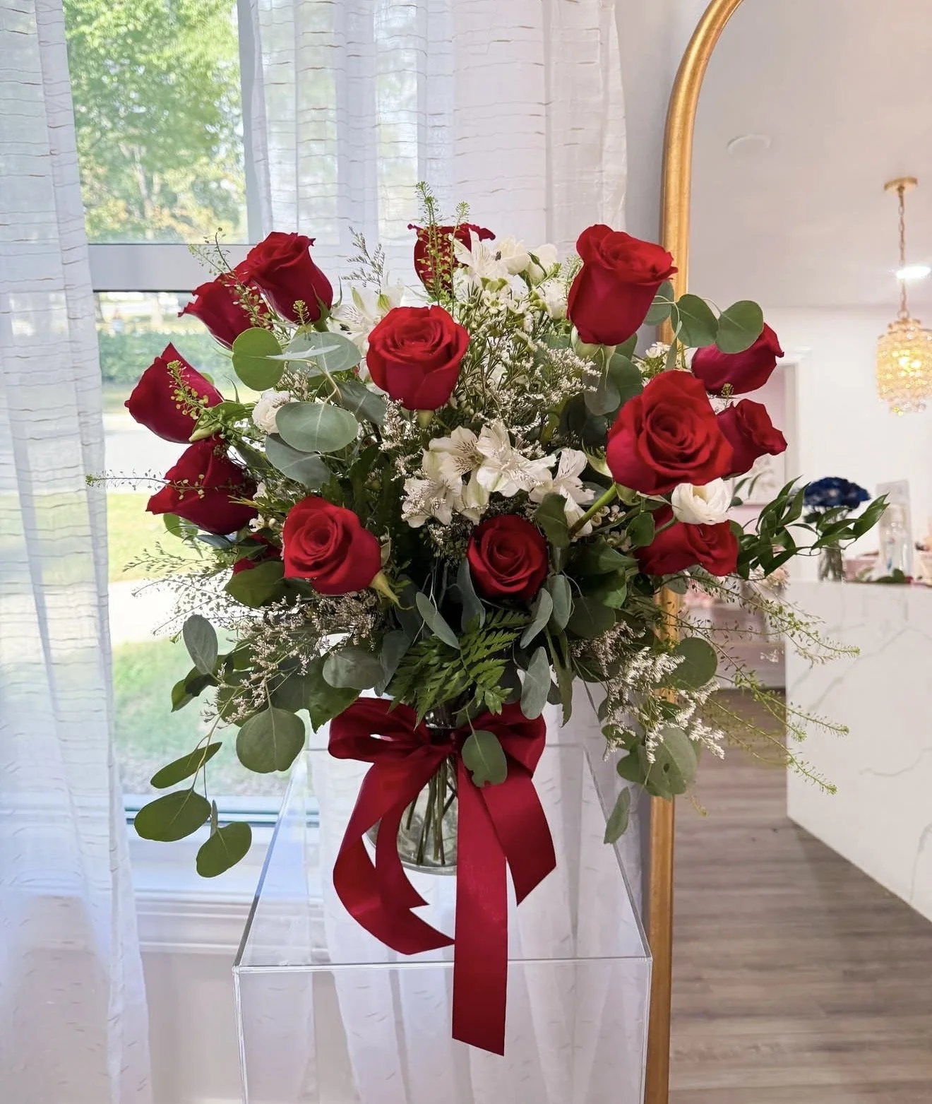 A bouquet of red roses, white lilies, and greenery in a glass vase with a red ribbon, placed on a clear stand indoors near a window with sheer curtains.