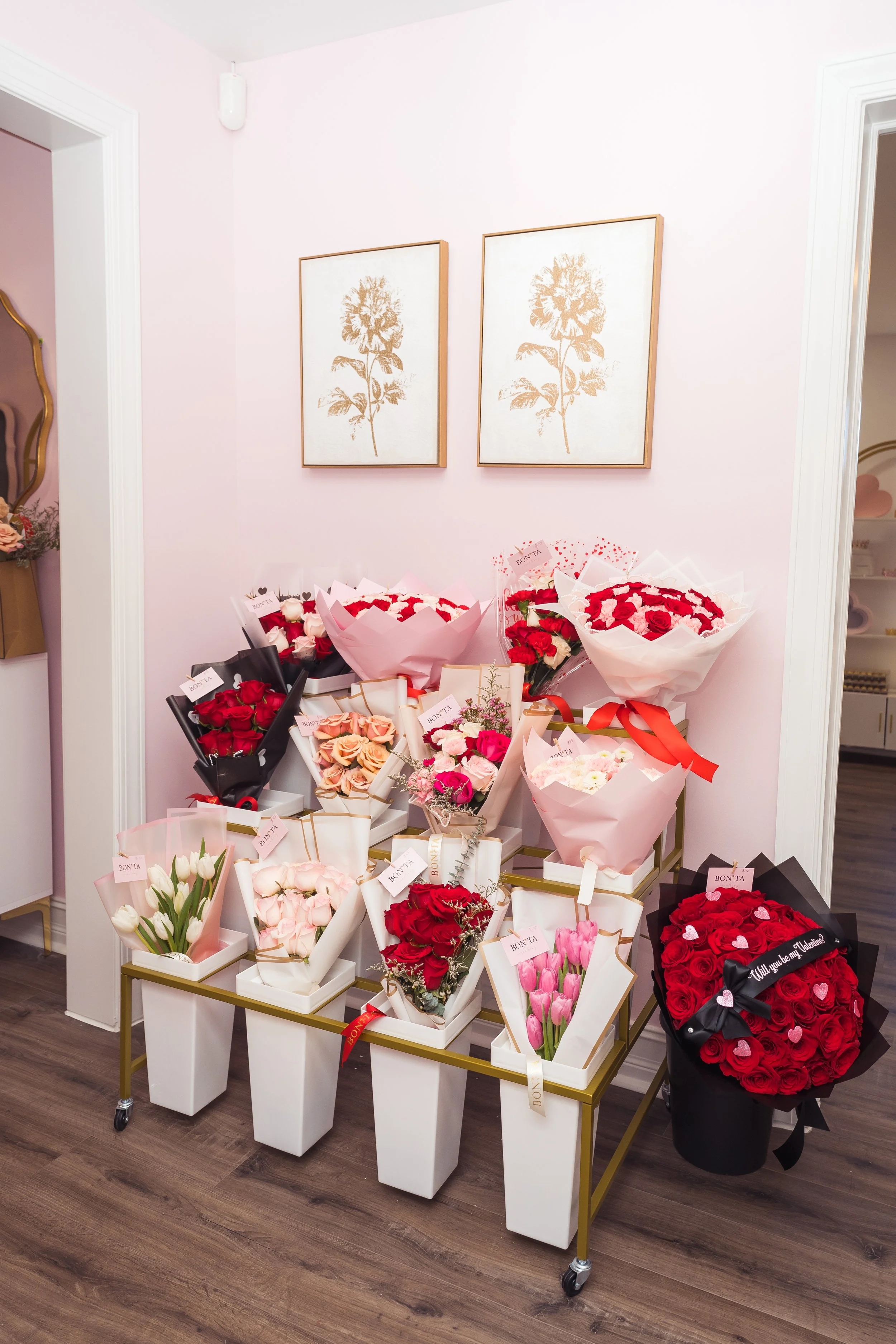 Display of various flower bouquets on a gold and white cart in a room with light pink walls and framed botanical prints.