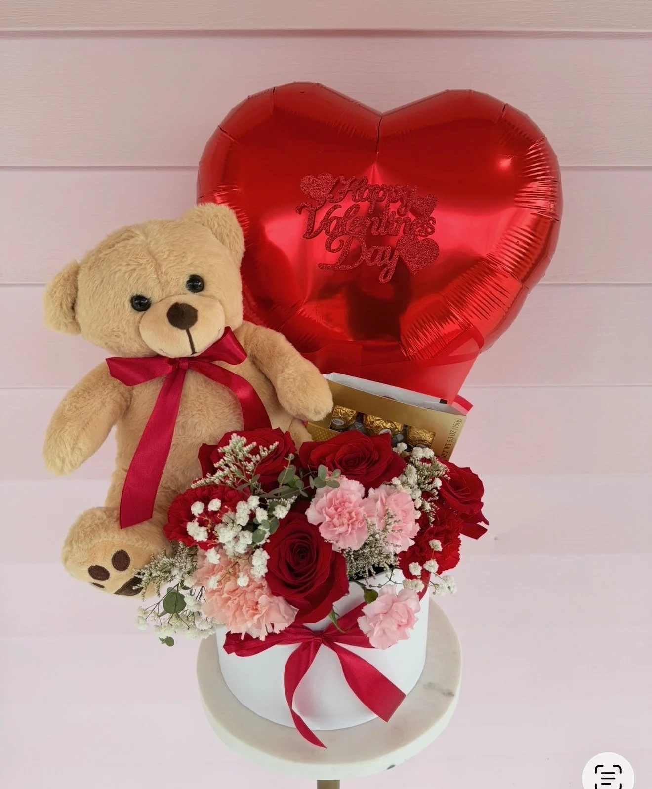 Valentine's Day gift arrangement with a teddy bear, red and pink flowers, a red heart-shaped balloon with "Happy Valentine's Day" written on it, and a box of chocolates, all on a white round table against a light pink wooden wall.