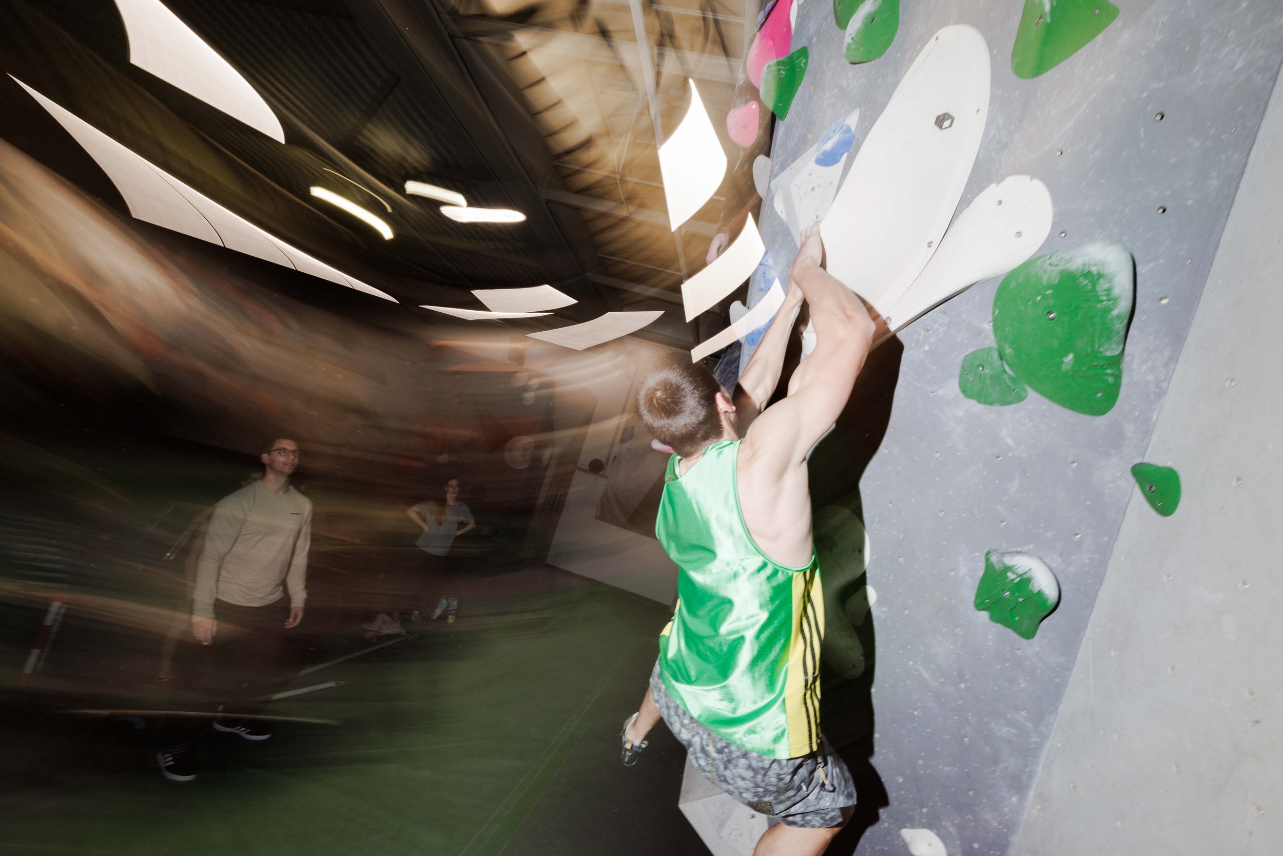 Ein junger männlicher Kletterer in einem grünen Tanktop, der an einer Kletterwand mit grünen und weißen Griffen hochklettert, während im Hintergrund einige Menschen zusehen.