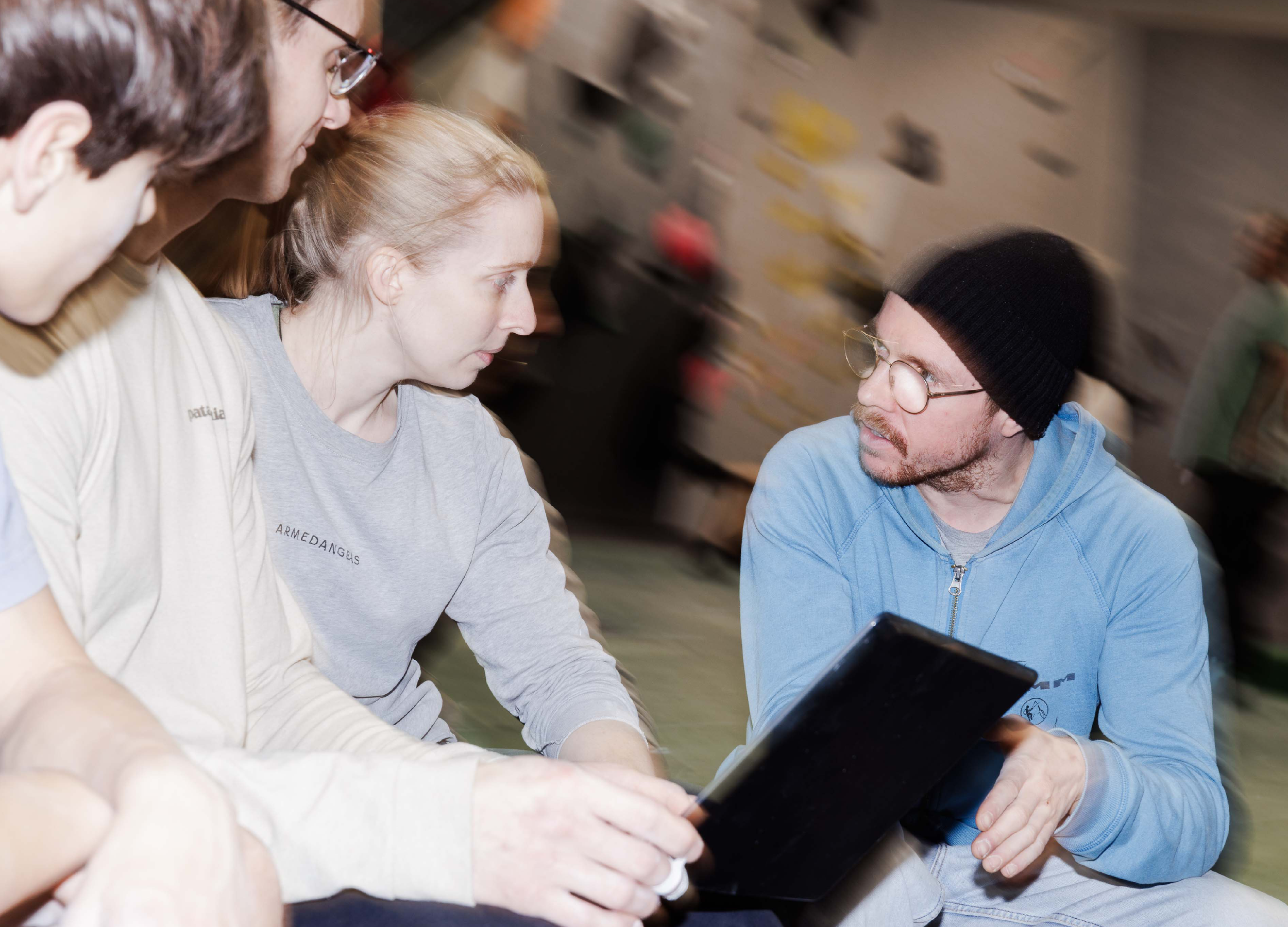 Drei junge Menschen, zwei Frauen und ein Mann, sitzen im Gespräch, wobei einer die Gruppe ein Tablet zeigt. Der Mann trägt eine schwarze Mütze, Brille und einen blauen Hoodie, die Frauen haben lange Haare, eine blond und eine braun, beide tragen graue Pullover.