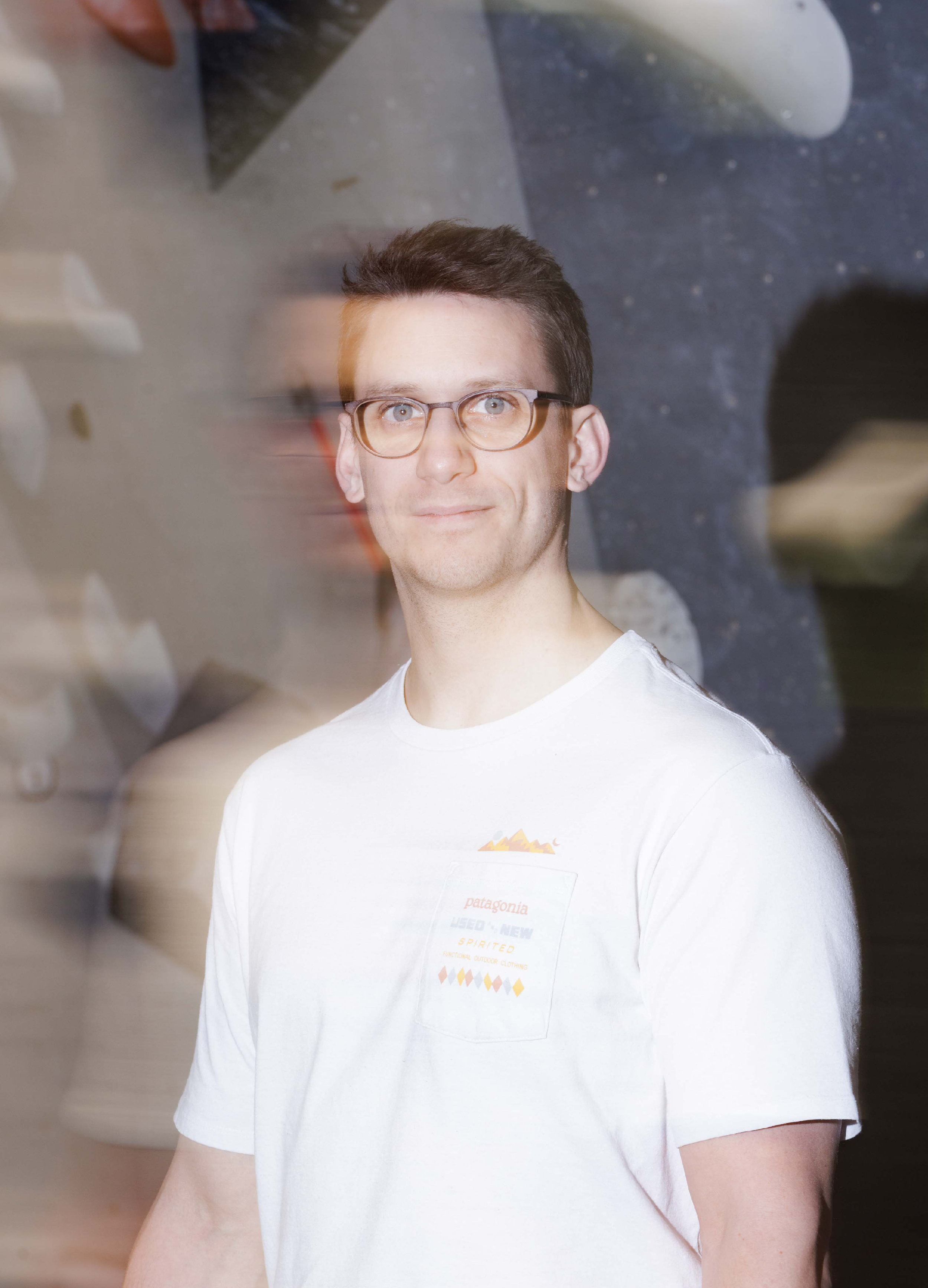 Junger Mann mit Brille steht vor dunklem Hintergrund, durch eine Glasfläche ist seine Spiegelung sichtbar, er trägt ein weißes T-Shirt mit Patagonian-Logo.