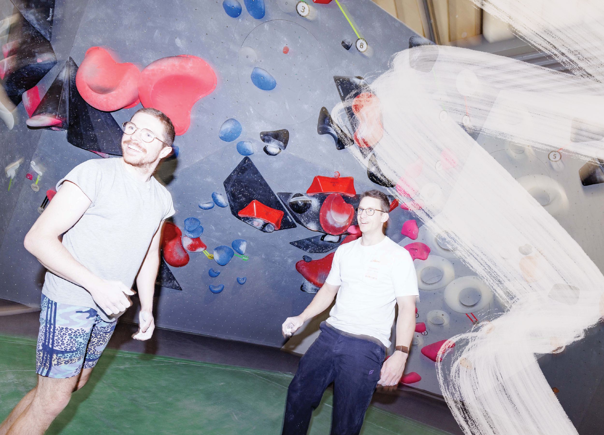 Zwei Männer in einem Indoor-Kletterzentrum, die vor einer Wand mit bunten Klettergriffen stehen und lachen.