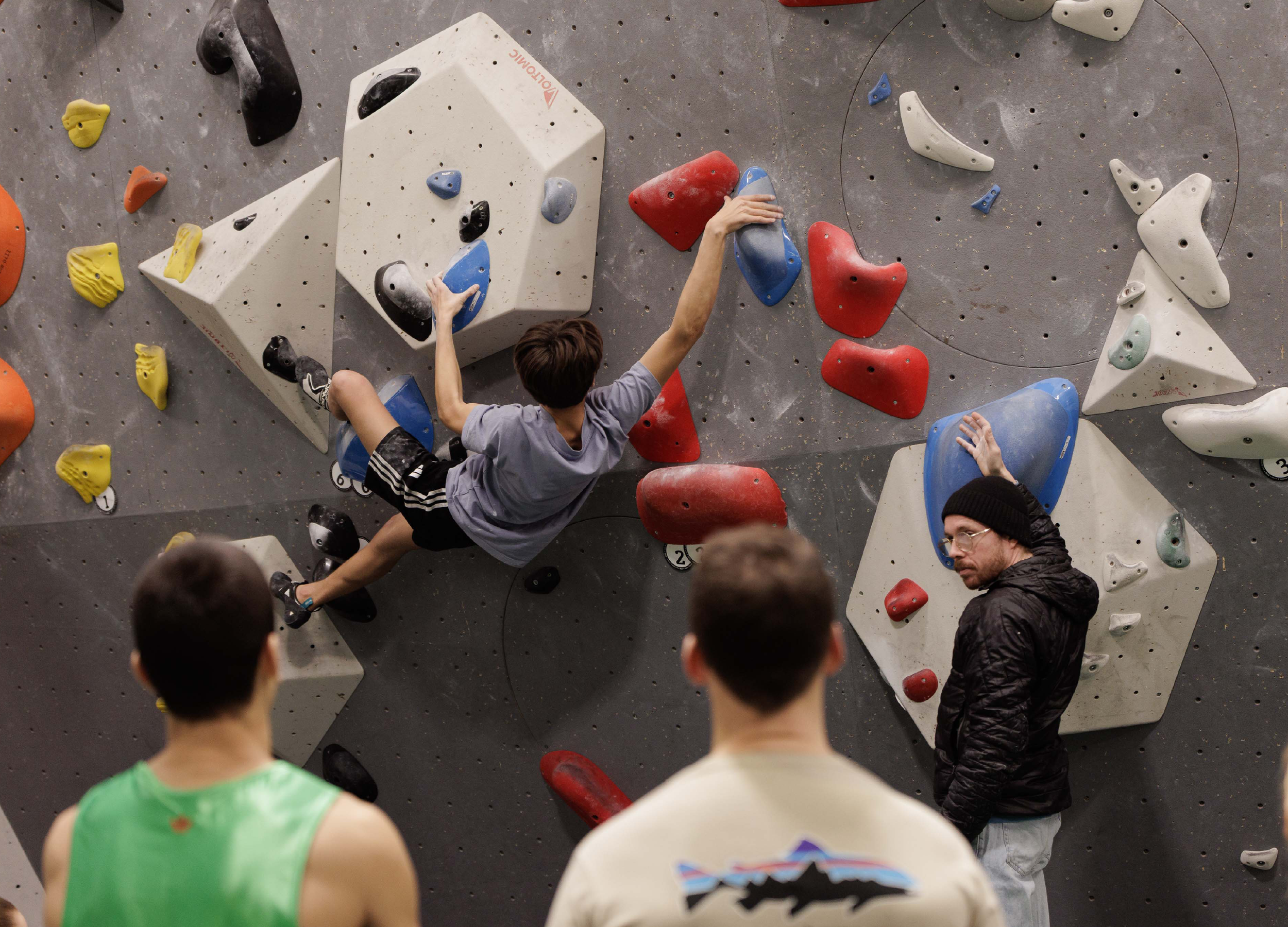 Junge Kletterer beim Bouldern an einer Kletterwand mit bunten Tritten, umgeben von drei Zuschauern in einer Kletterhalle.