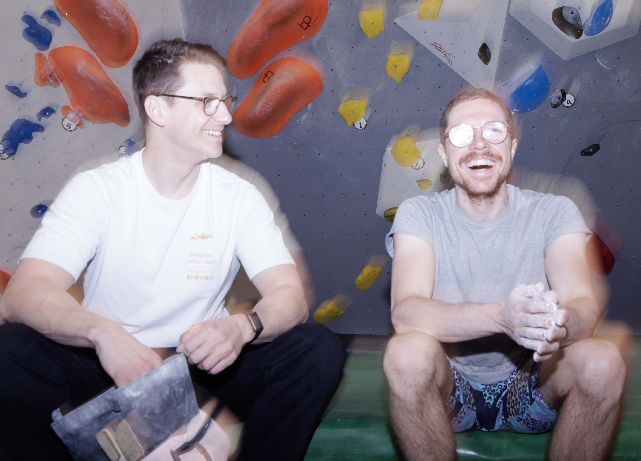 Zwei Männer sind in einer Kletterhalle mit bunten Klettergriffen an der Wand. Sie lachen und scheinen eine Pause zu machen.