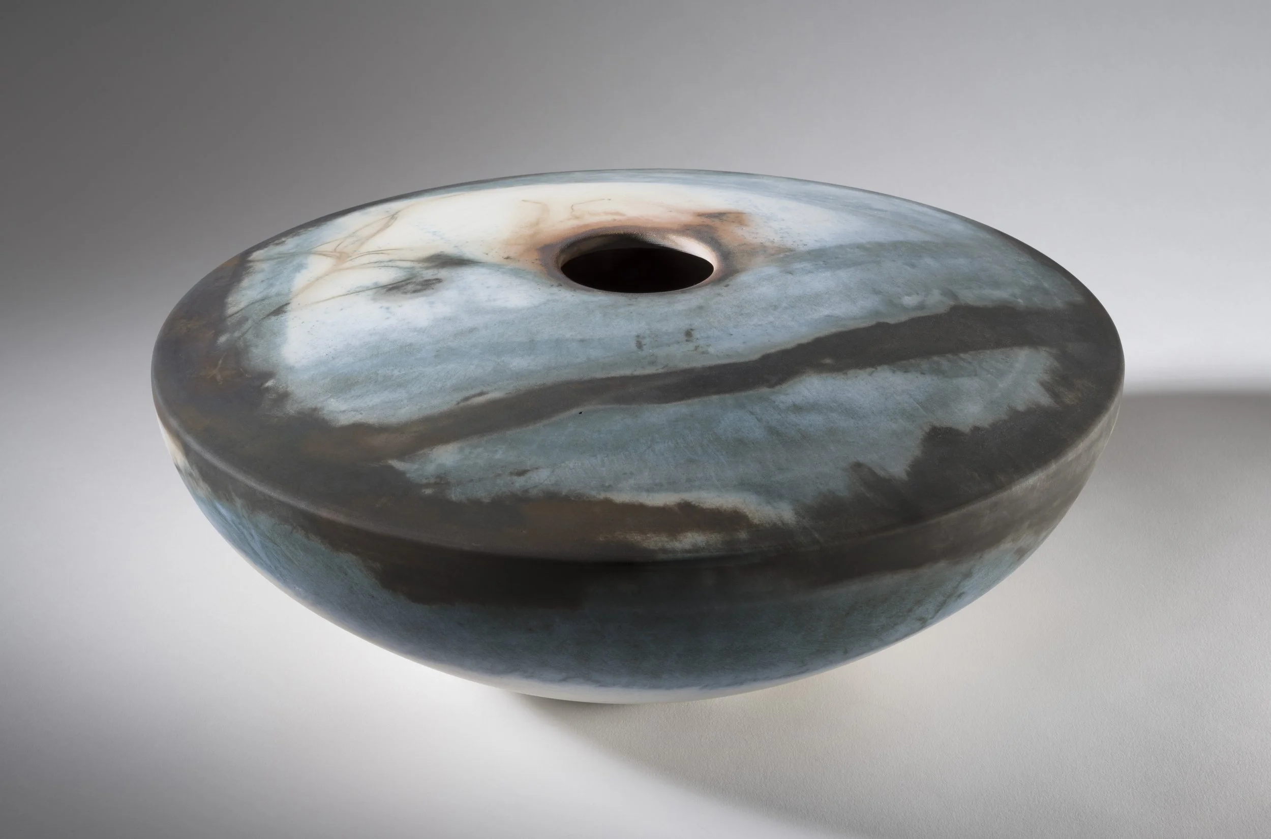 A round, convex sculptural vessel with a hole in the center, featuring swirling blue, gray, and white patterns.