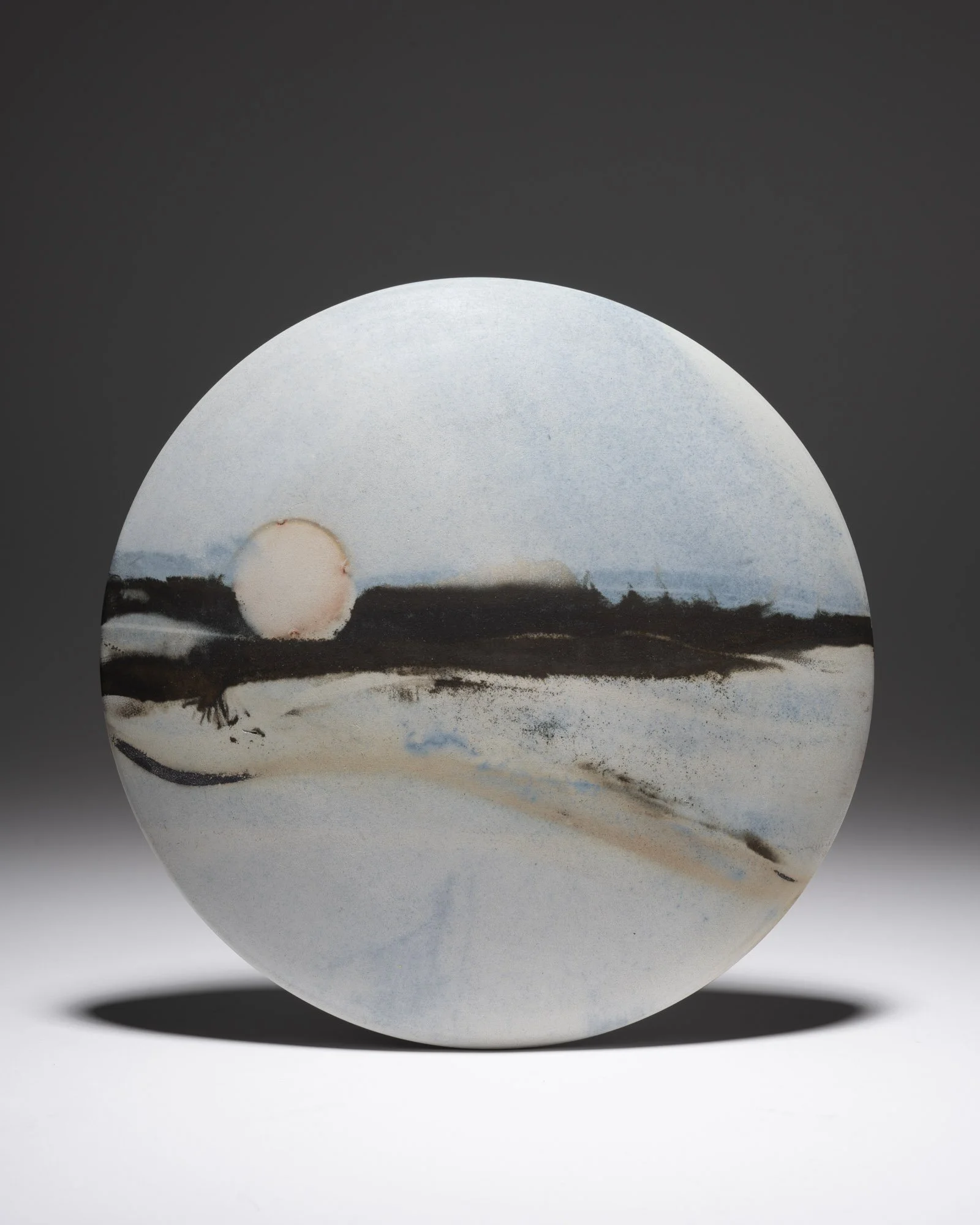 A circular ceramic decorative wall disc, with an abstract landscape featuring a dark shoreline, pale sky, and a large pinkish sun near the horizon.