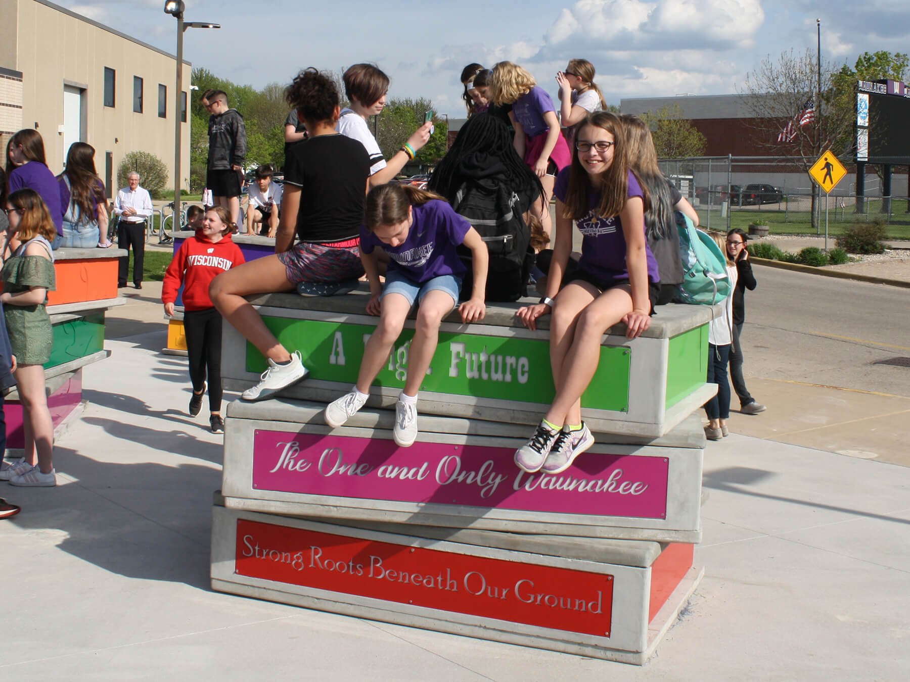 Group of students sitting and standing on a colorful outdoor art installation with message about roots and education and community in a school yard.