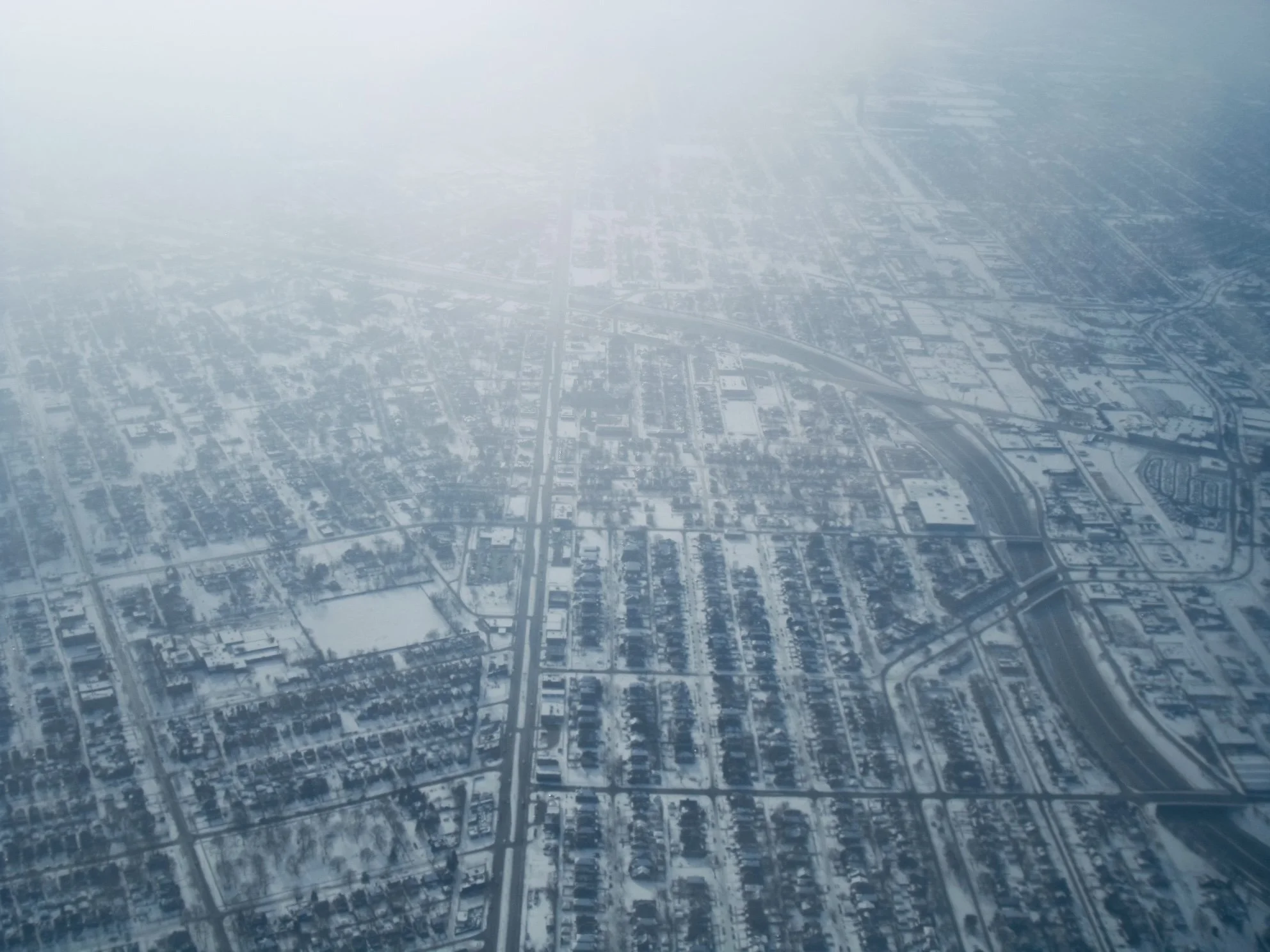 Aerial view of a snow-covered city grid with roads, buildings, and a highway overpass, shrouded in fog or haze.