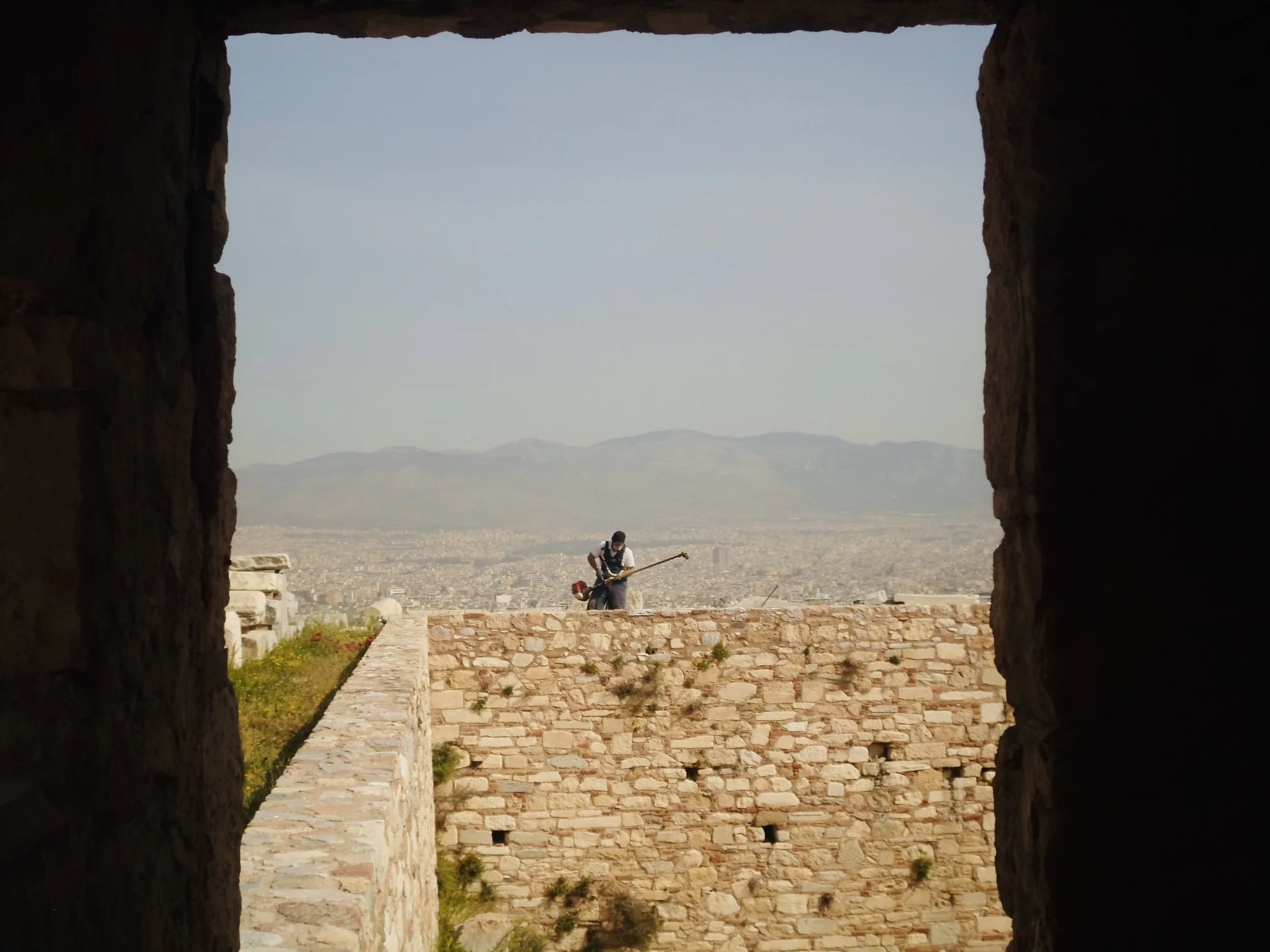A person standing on a stone wall, playing a guitar, viewed through a window in an ancient stone structure, overlooking a city and mountains in the distance.