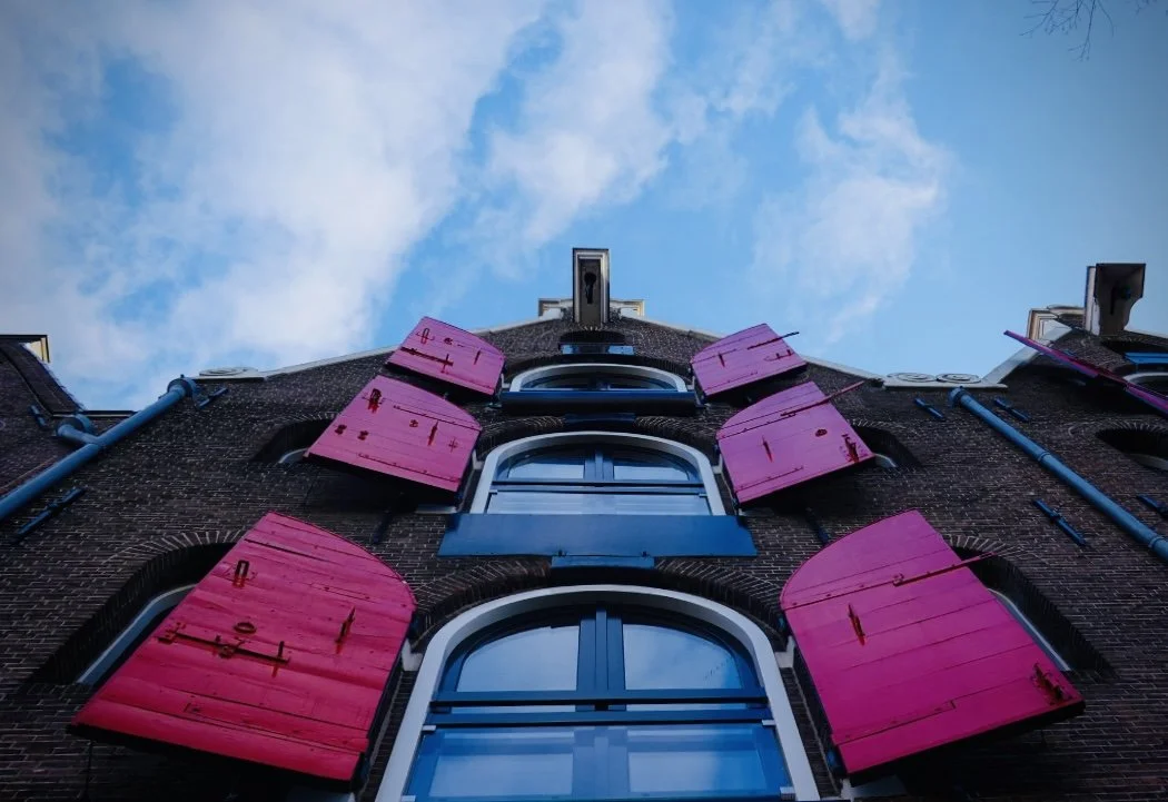 Upward view of a brick building with three windows and pink shutters, with a blue sky and clouds in the background.