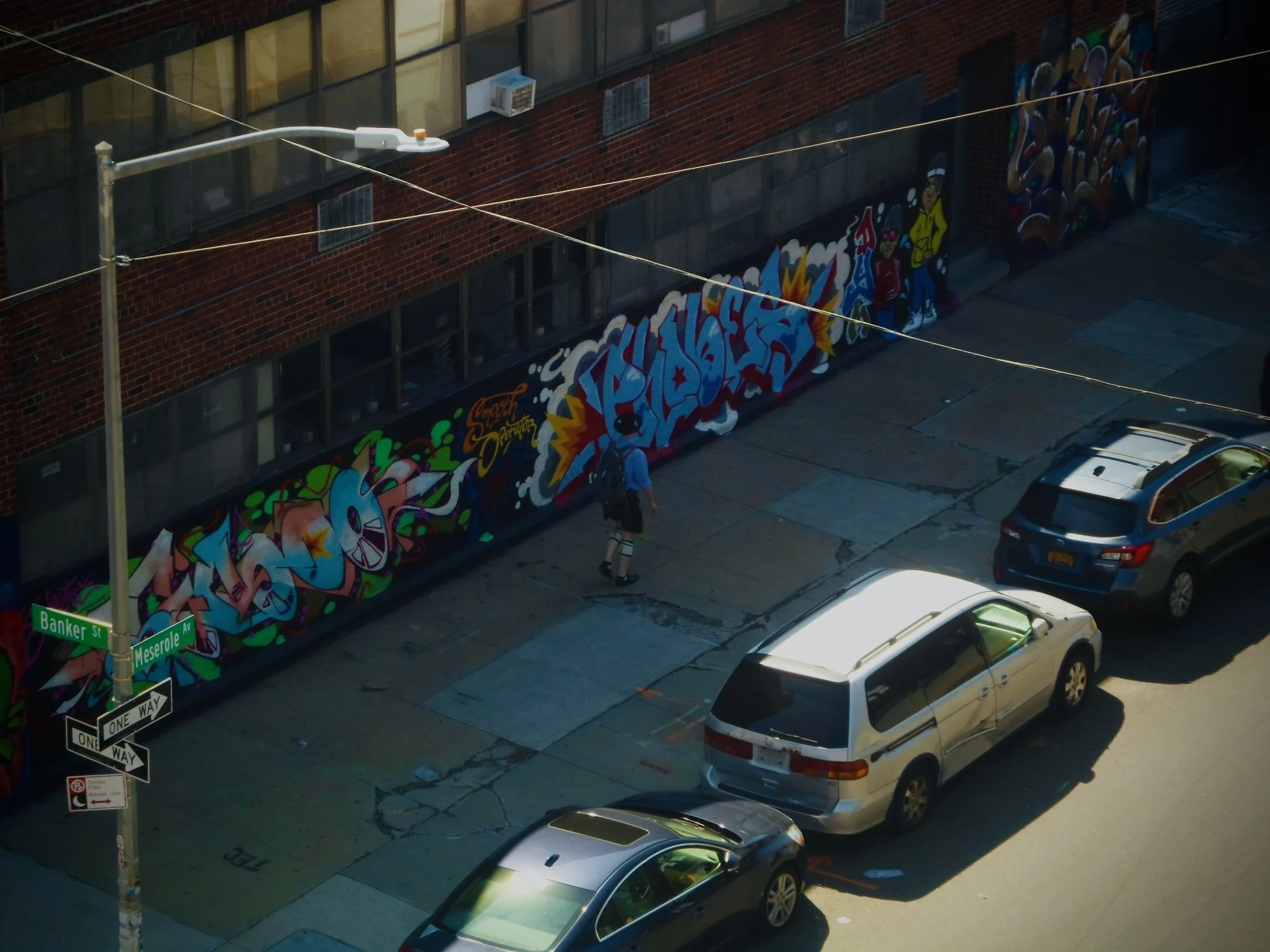 Street view of a brick building with large graffiti art on the wall, street signs indicating Banker Street and Meserole Avenue, parked cars, a streetlight, and a person walking on the sidewalk.