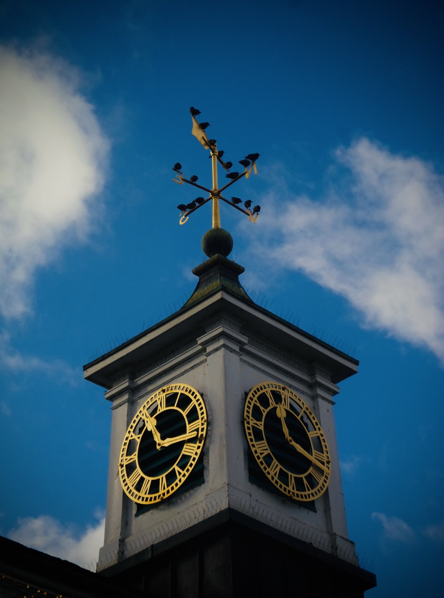 A clock tower with two clocks displaying approximately 11:02, topped with a weather vane with birds on it, against a partly cloudy sky.