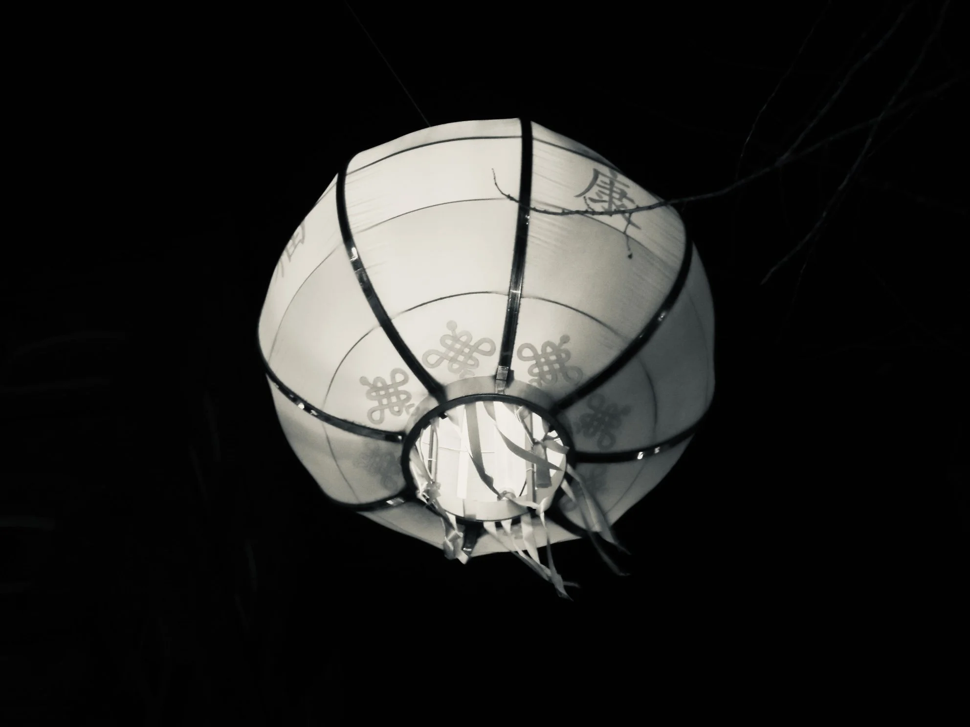 A glowing white paper lantern with black metal frame details hanging in the dark, with some wires and small branch twigs surrounding it.